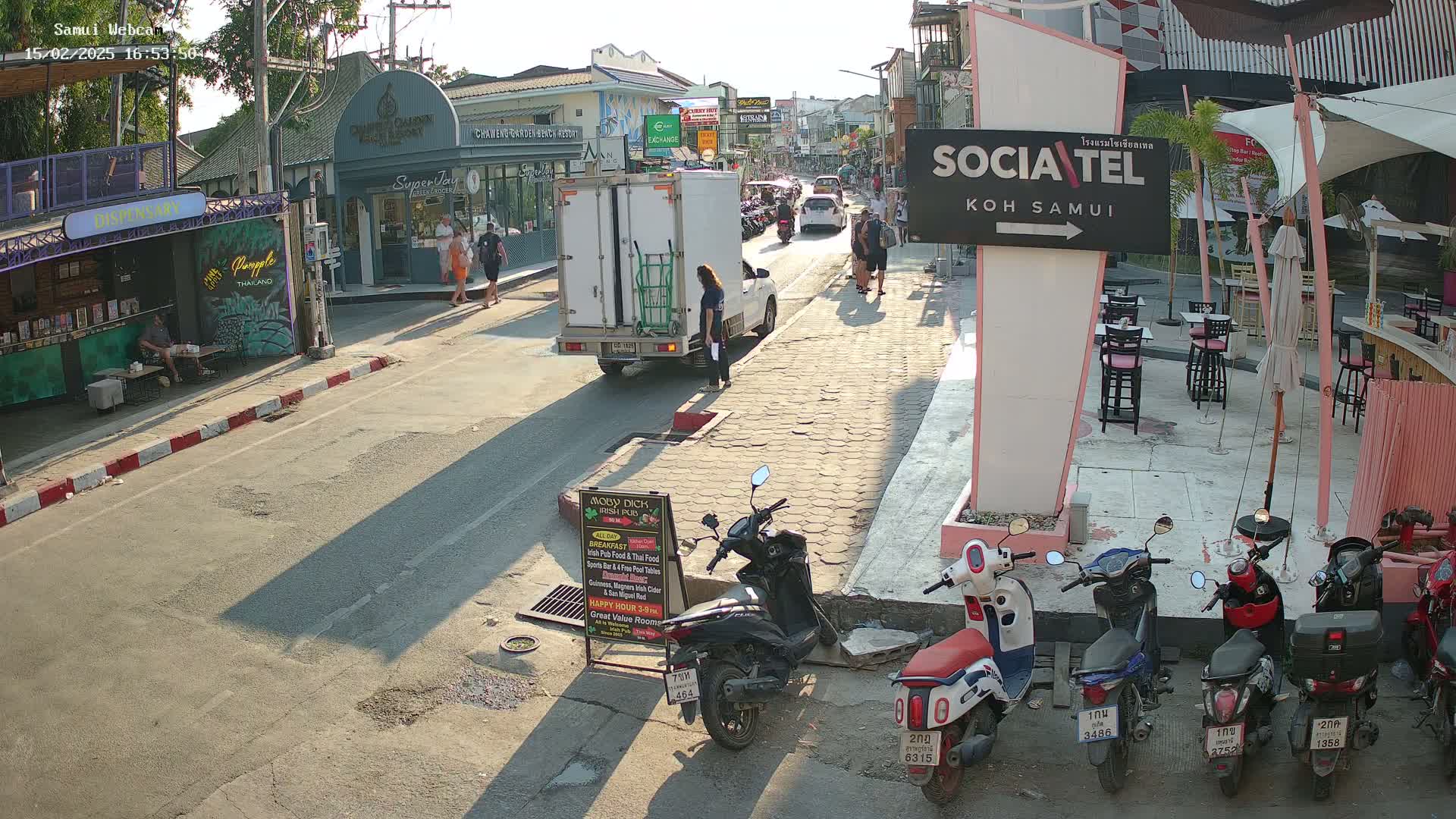 A sunny street scene shows pedestrians walking past shops and restaurants, with several parked motor scooters and a white delivery truck on a paved road.