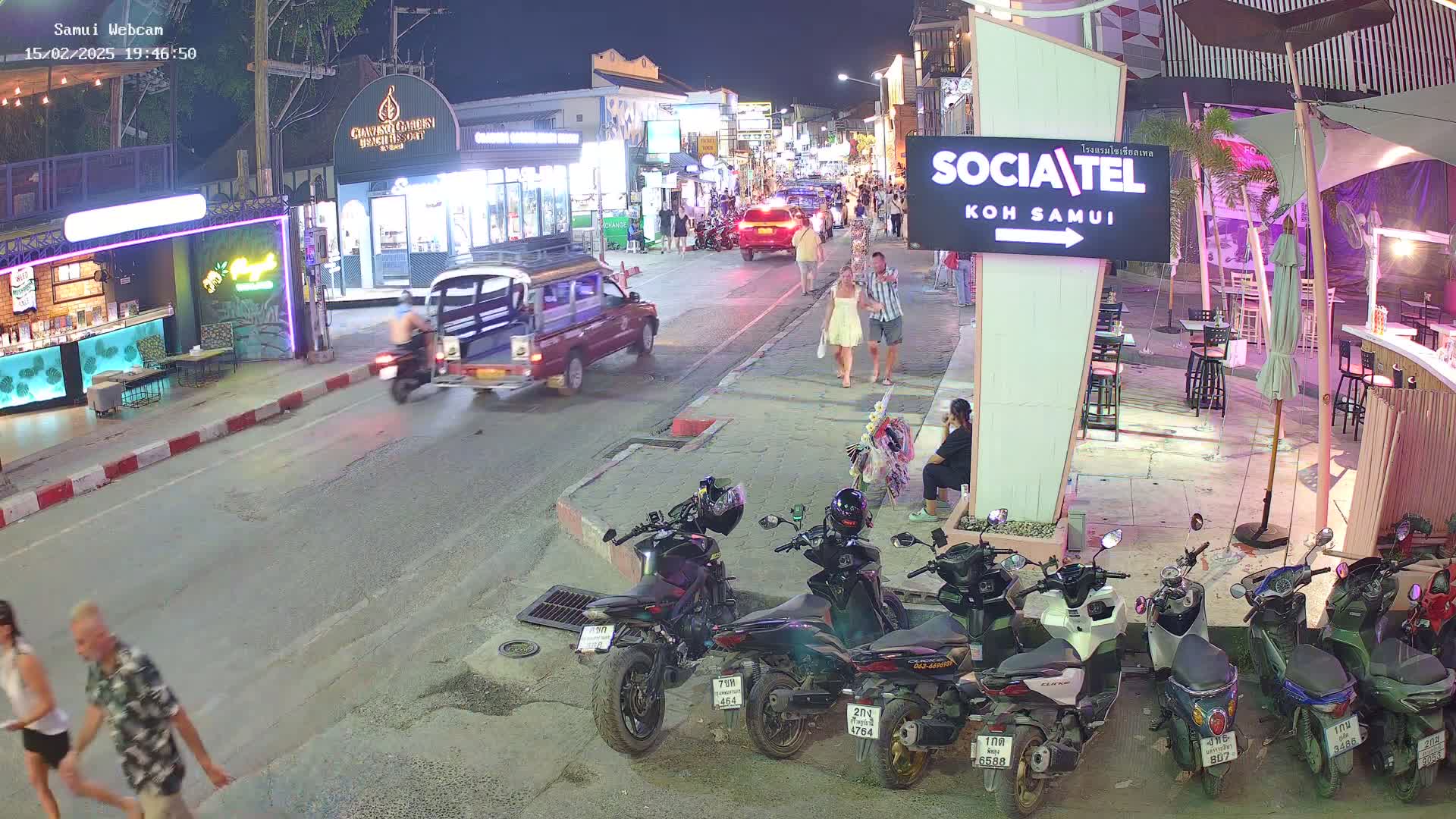 A nighttime street scene shows pedestrians and vehicles, including motorcycles parked along the curb and a pickup truck driving down the road, under clear skies.