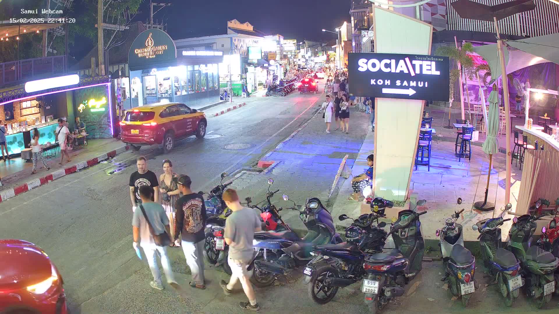 A nighttime street scene shows a group of people standing together near parked motorcycles and scooters, with other pedestrians and vehicles further down the road under the lights of shops and restaurants.