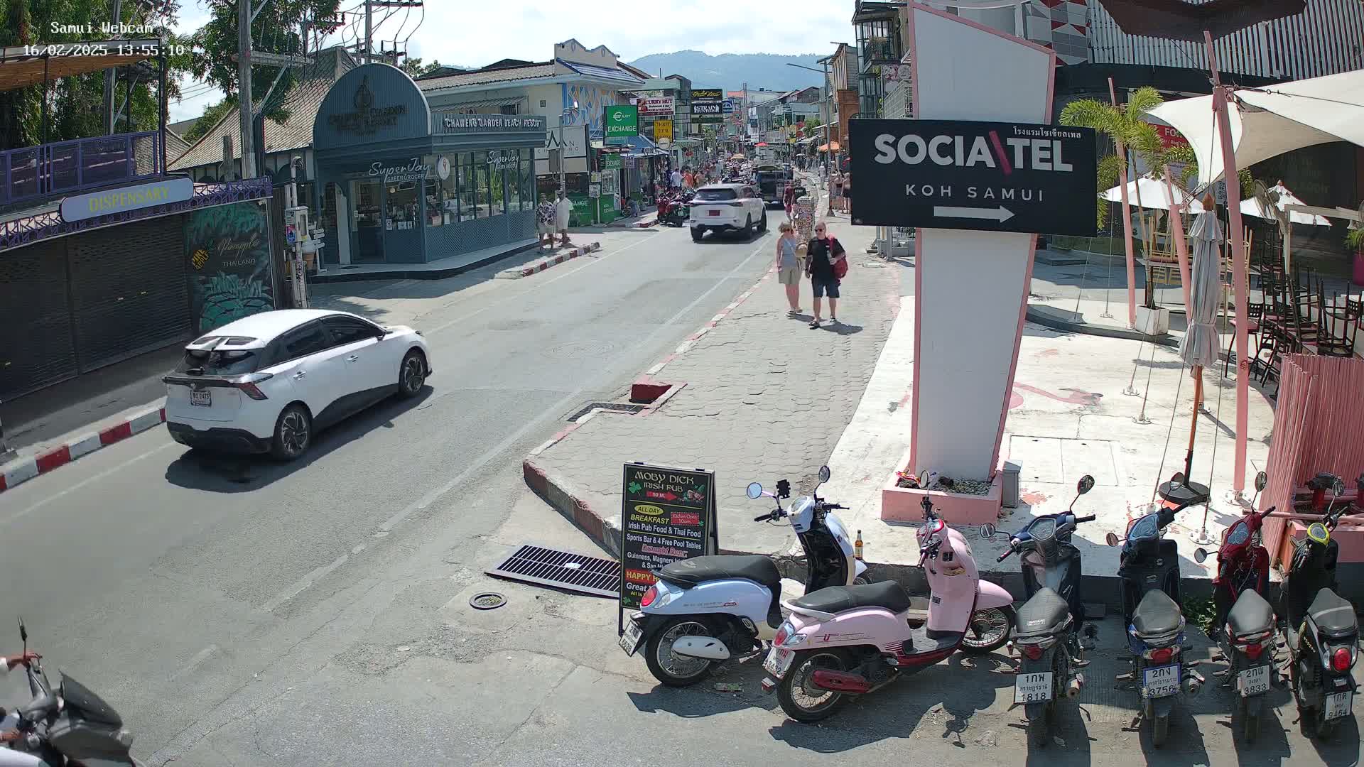A sunny street scene shows a white car driving away from the camera, several parked motorbikes, and pedestrians walking past shops and restaurants.