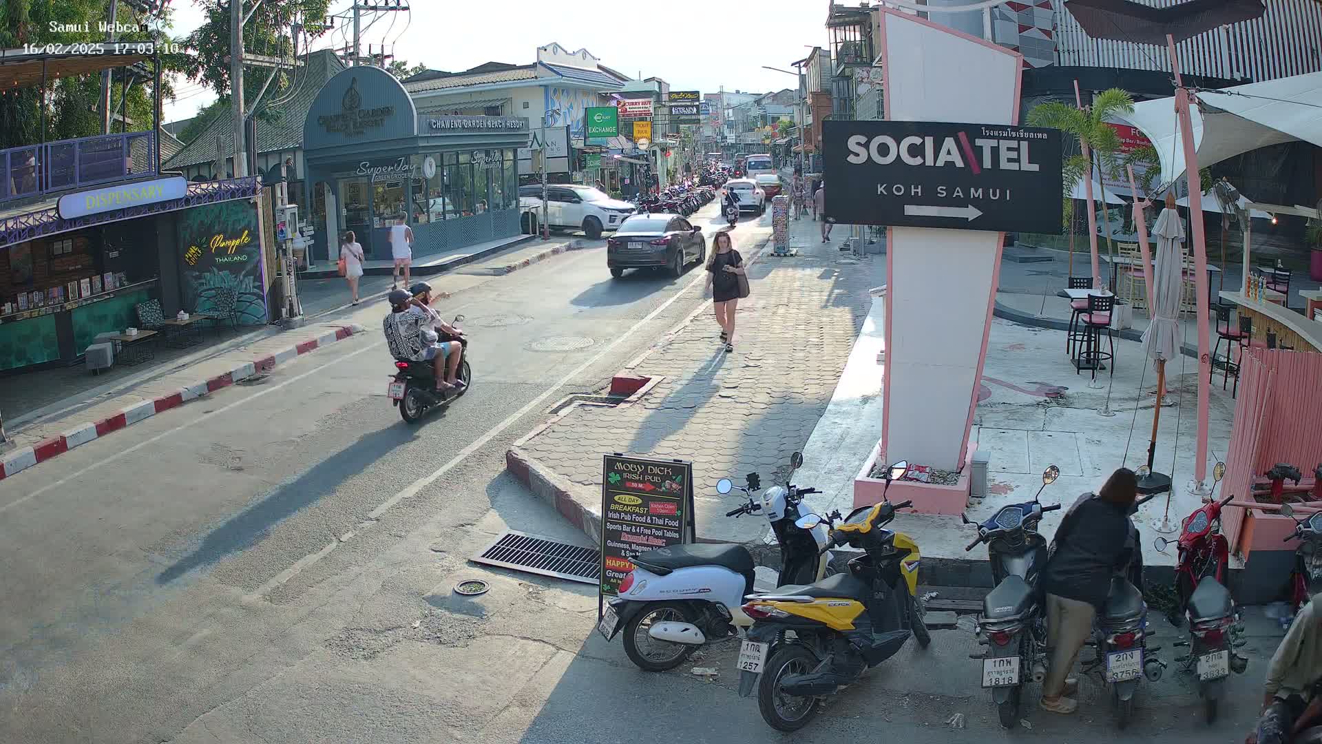 A sunny street scene shows people walking and riding motorbikes past shops and restaurants, with several parked motorbikes near a pink building.
