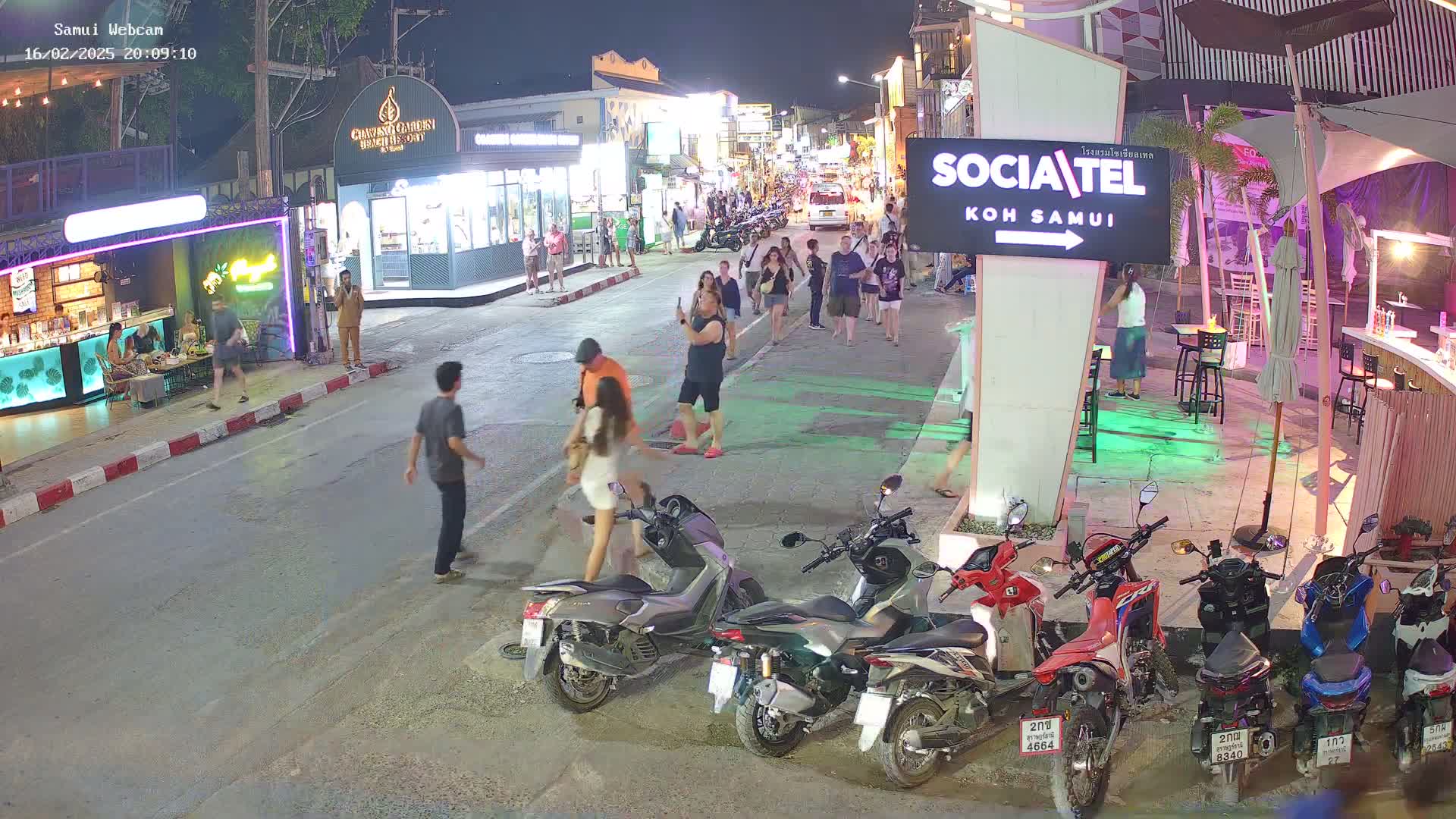 A nighttime street scene shows pedestrians walking past shops and restaurants, with several parked motorcycles and scooters lining the side of the road.
