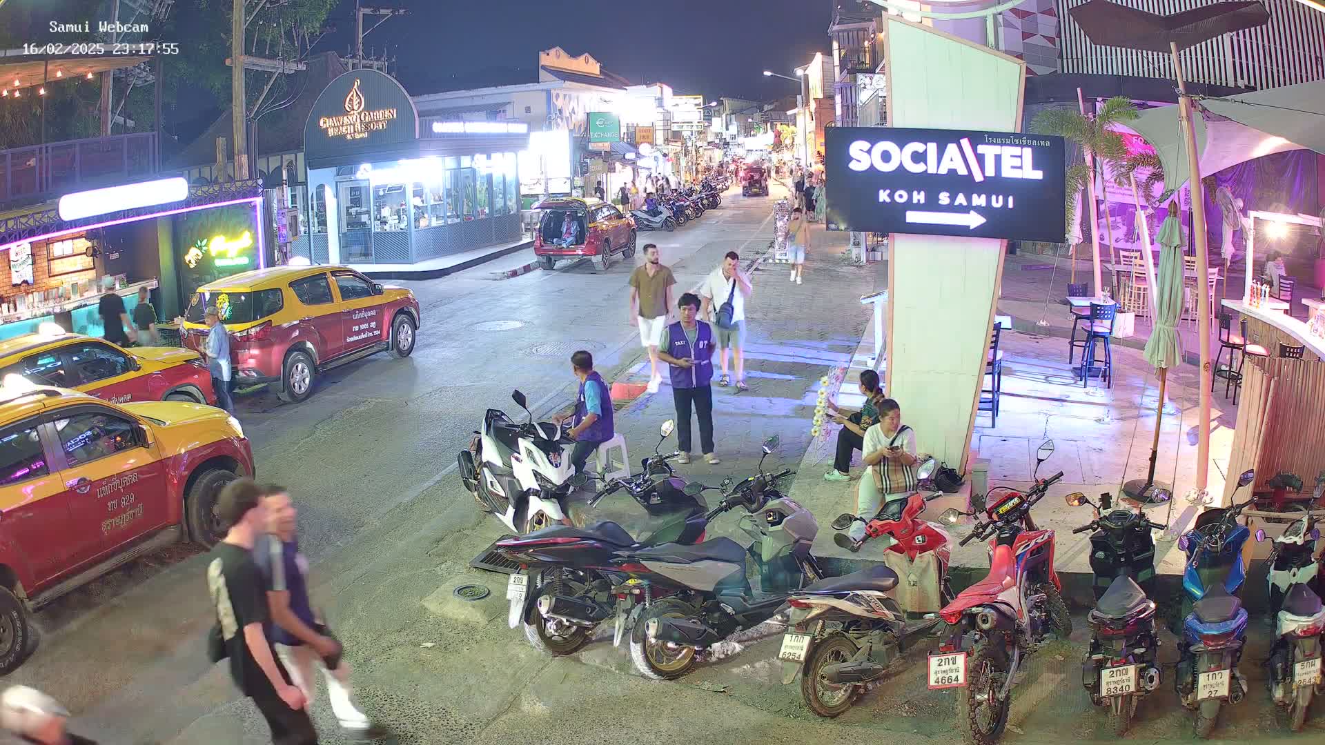 At night, several people stroll along a street lined with parked vehicles and businesses, under a clear sky.