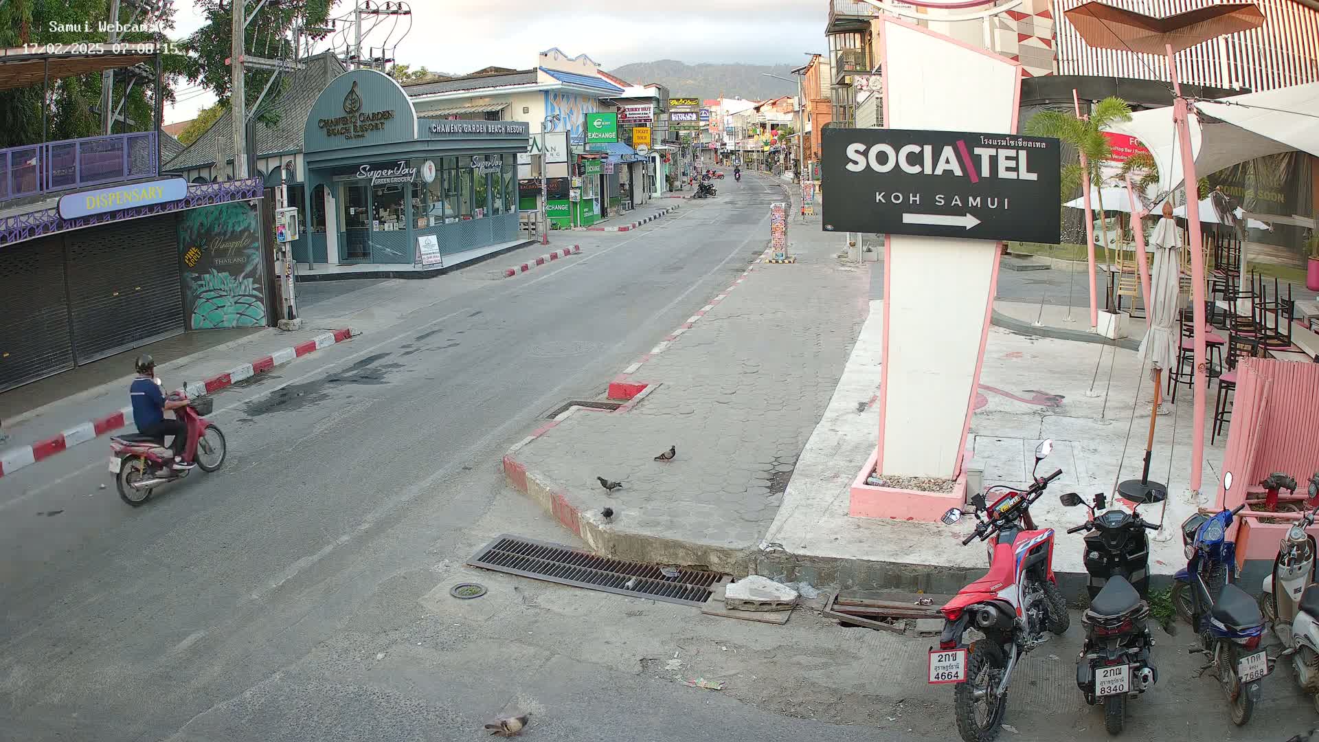 A mostly empty street scene in bright daylight shows several shops and restaurants along the sides of the road, with a few motorcycles parked and one person riding a scooter.