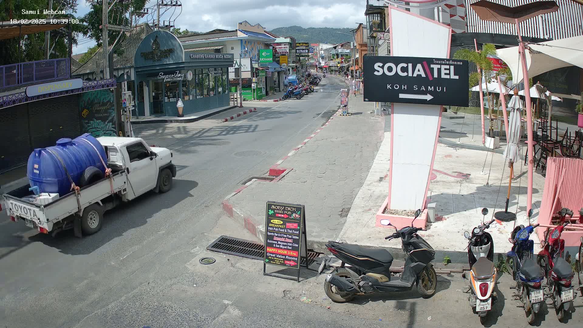 A sunny street scene shows a white pickup truck carrying a large blue water tank, several parked motorbikes, and shops and restaurants along the road.