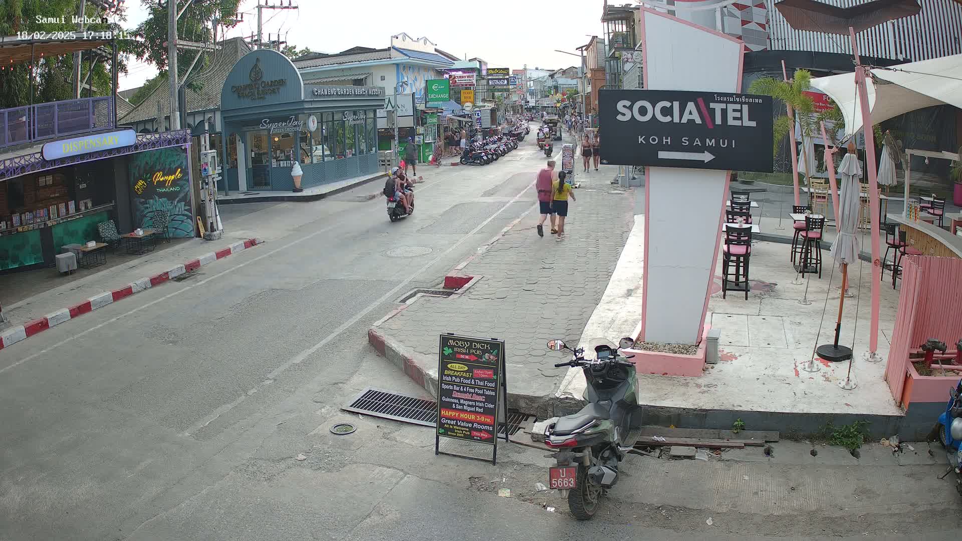 A street scene on a partly cloudy day shows pedestrians and motorbikes on a road lined with shops and outdoor seating areas.