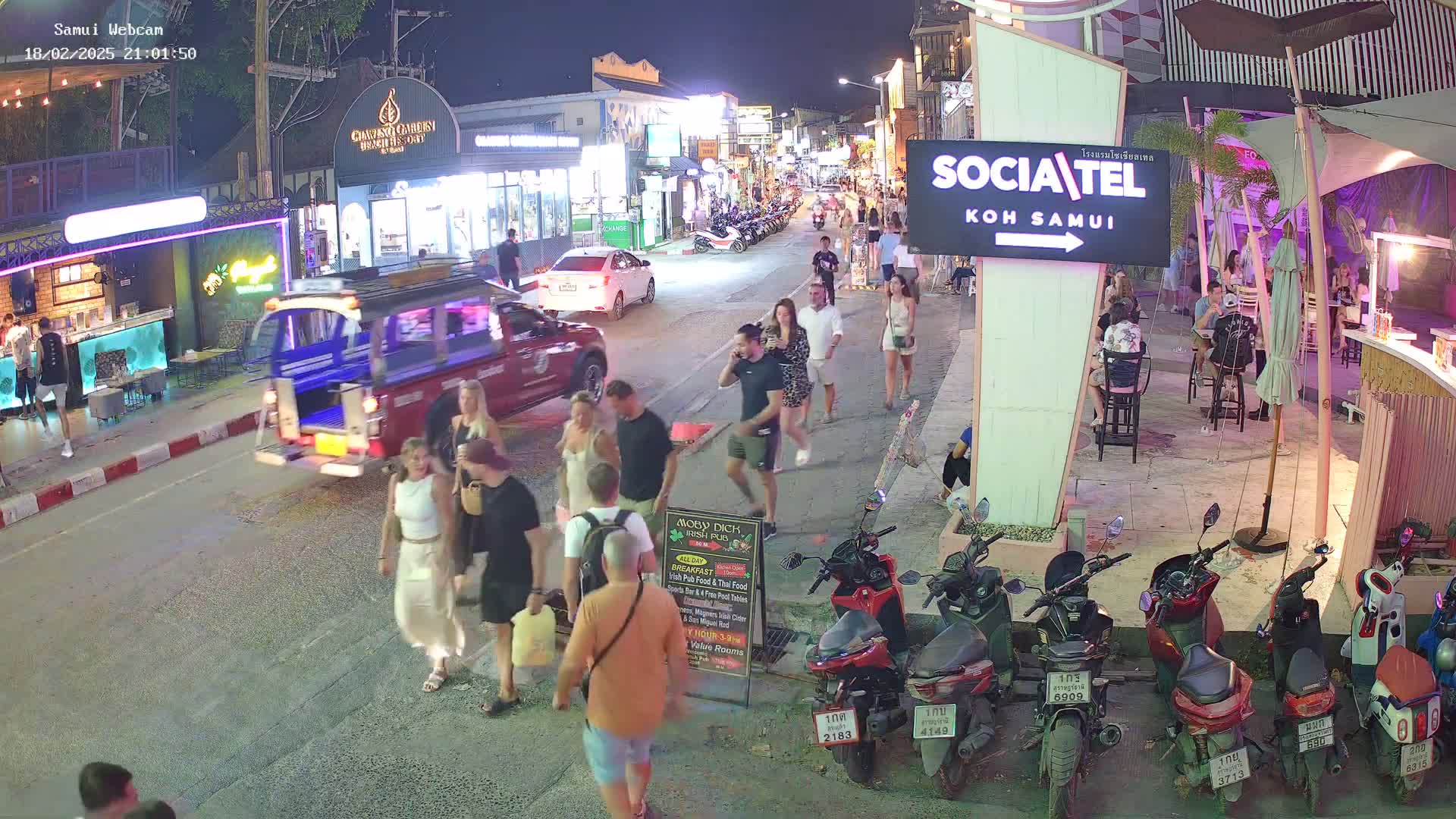A nighttime street scene shows pedestrians walking among parked motorbikes and passing vehicles, near brightly lit shops and restaurants.