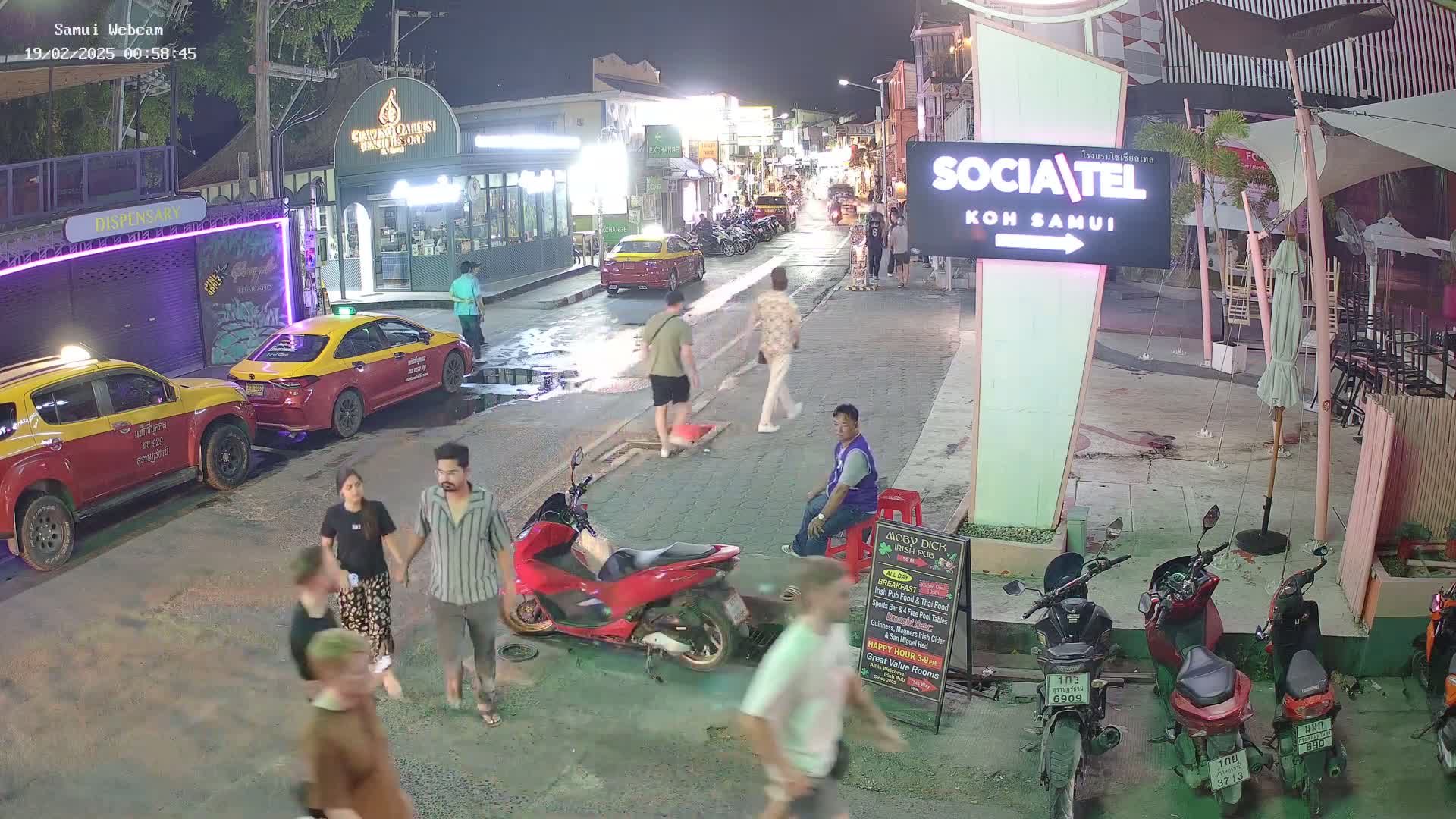 A nighttime street scene shows several people walking and standing amidst parked cars and motorcycles, under clear skies.