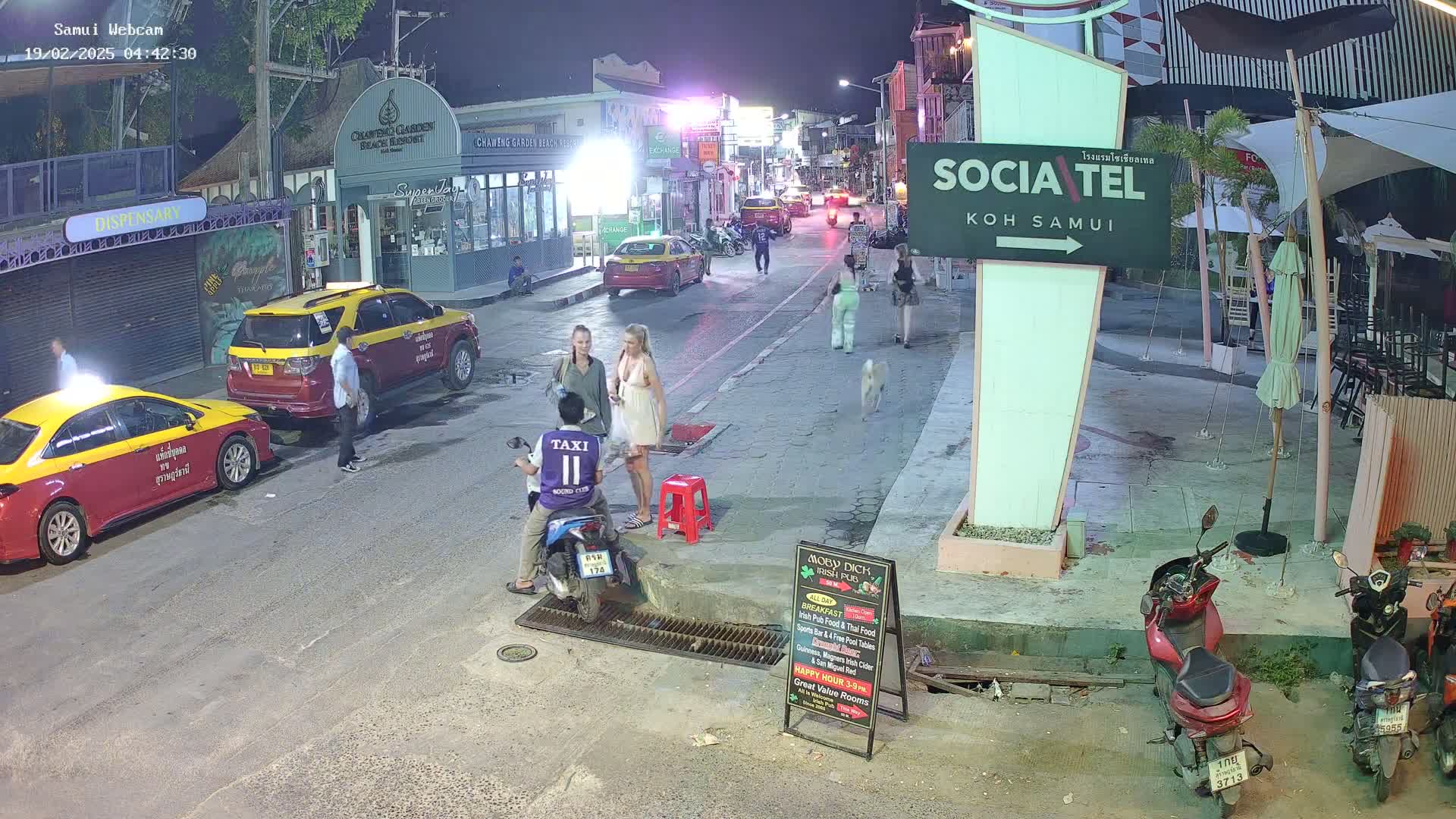 At night, several people are walking and standing on a street lined with buildings, while two women converse near a motorcyclist and several parked taxis.