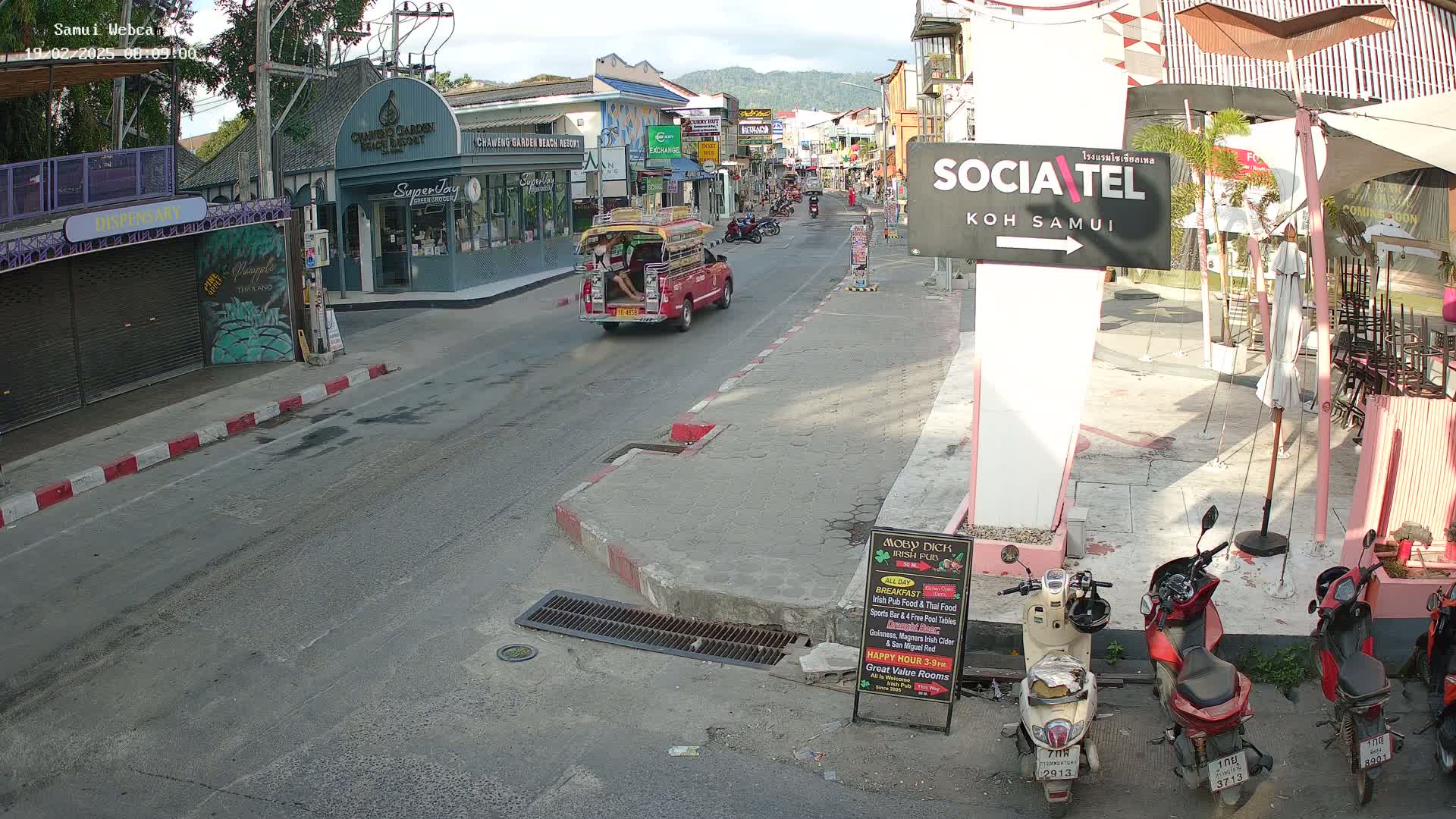 A sunny street scene shows a red songthaew driving away from the camera, several parked scooters, and various businesses along a street lined with buildings.