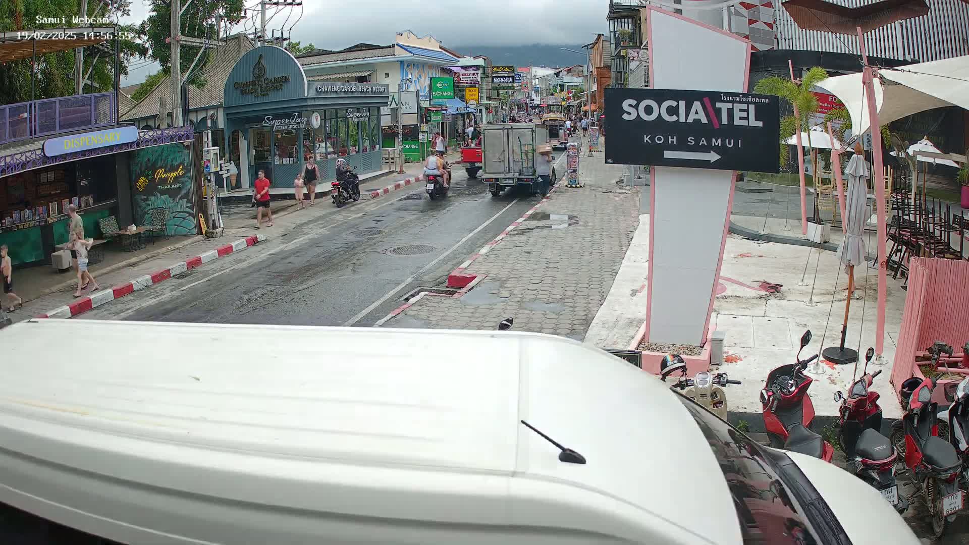 A wet street scene shows pedestrians and motorbikes on a roadway lined with shops and restaurants under an overcast sky.