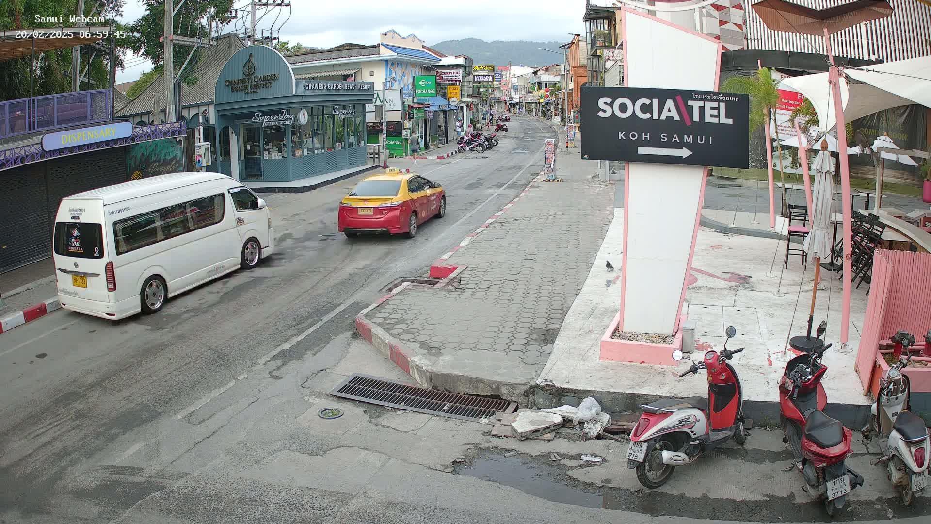 A street scene shows a white van and a yellow taxi driving past shops and restaurants under an overcast sky, with several parked motor scooters nearby.