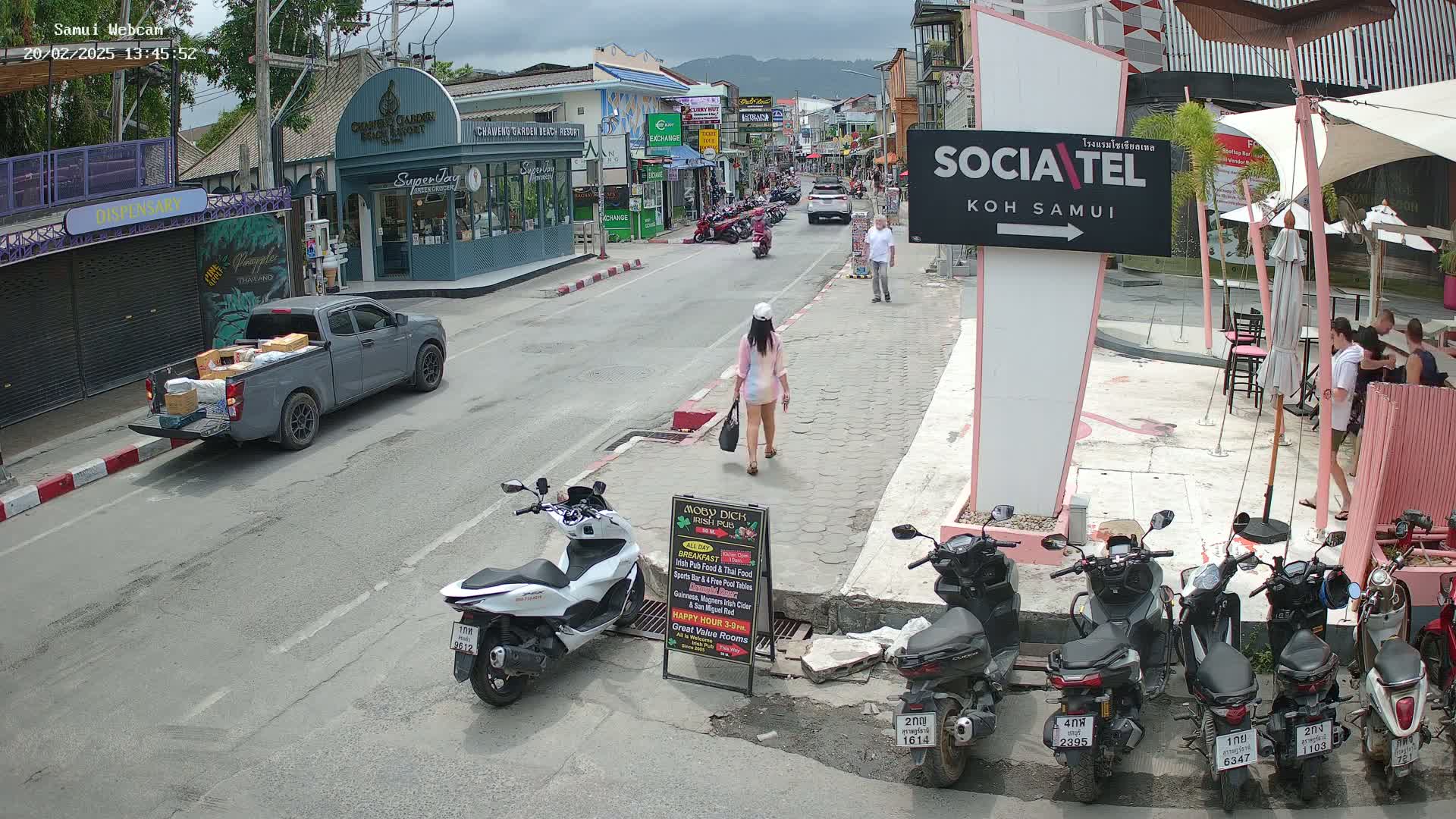 A street scene on a cloudy day shows a woman walking, parked motorcycles, a pickup truck carrying boxes, and people walking and sitting at outdoor cafes.