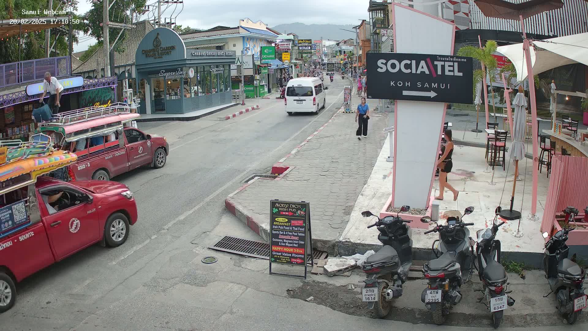 A street scene shows several red pickup trucks, motorbikes parked along the side of a road, and pedestrians walking past shops and restaurants on a partly cloudy day.