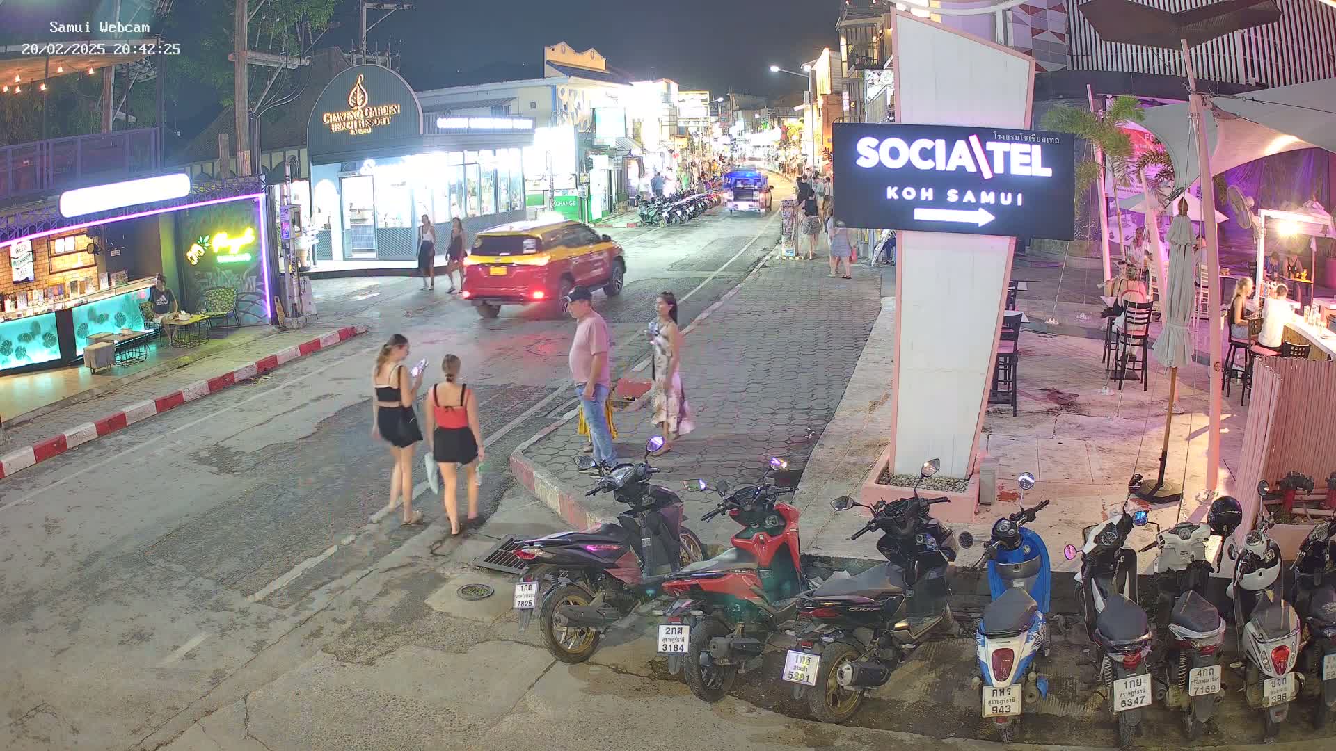 A nighttime street scene shows pedestrians and parked motorbikes along a road lined with shops and restaurants, under clear skies.