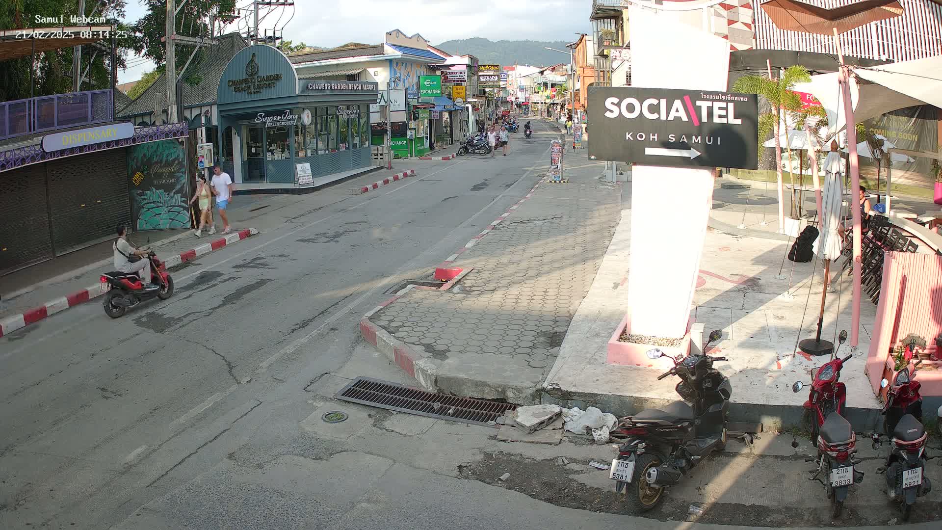 A sunny street scene shows a lightly trafficked road with parked and moving motorbikes, pedestrians, and shops lining both sides under a partly cloudy sky.