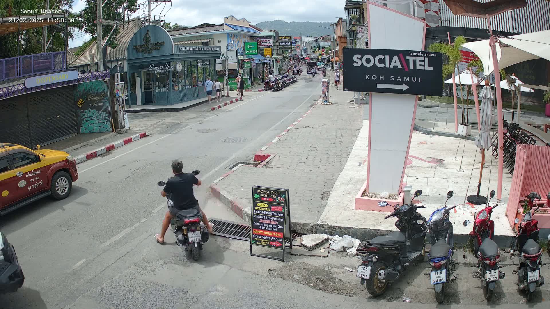 A man on a scooter drives down a street lined with shops and parked motorcycles under a partly cloudy sky.