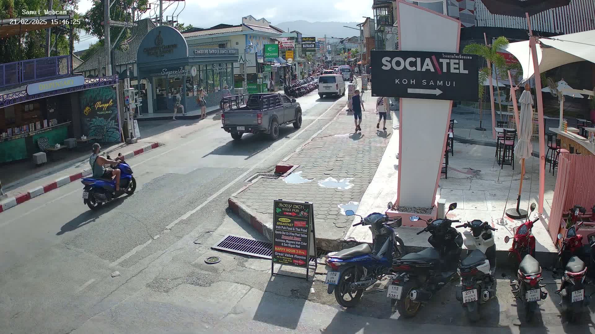 A sunny street scene shows a man on a blue scooter, parked motorbikes, and pedestrians walking past shops and restaurants.