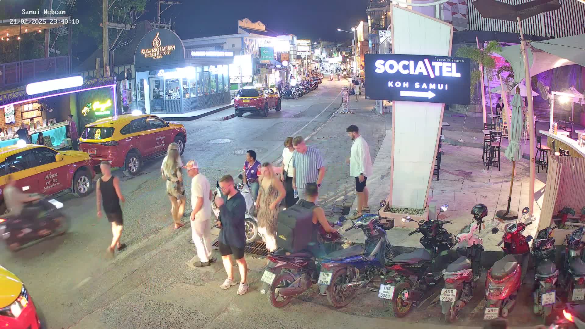A nighttime street scene shows people walking and standing near parked cars and motorbikes under outdoor lighting.