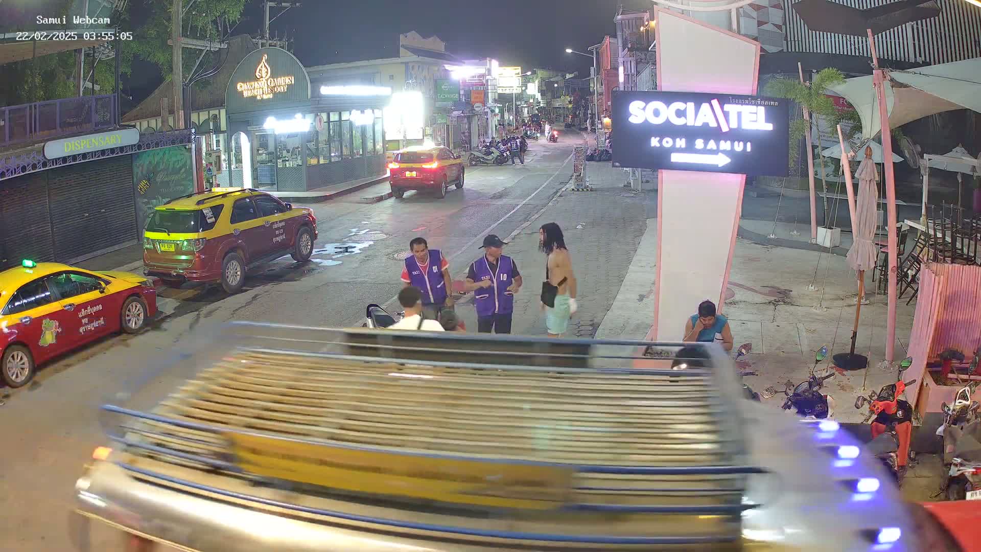At night, several people stand on a street in front of shops and restaurants, with parked cars and motorcycles nearby.