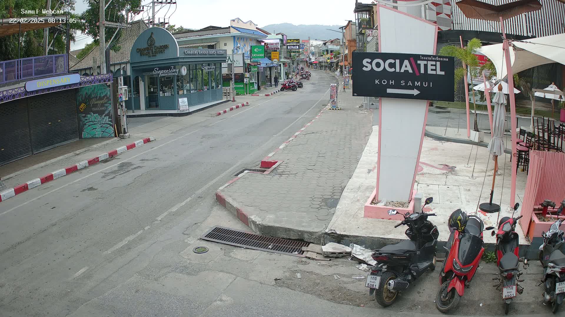 A mostly empty street scene on a cloudy day shows several parked motorbikes, shops, and buildings along the road, with a few pedestrians visible in the distance.