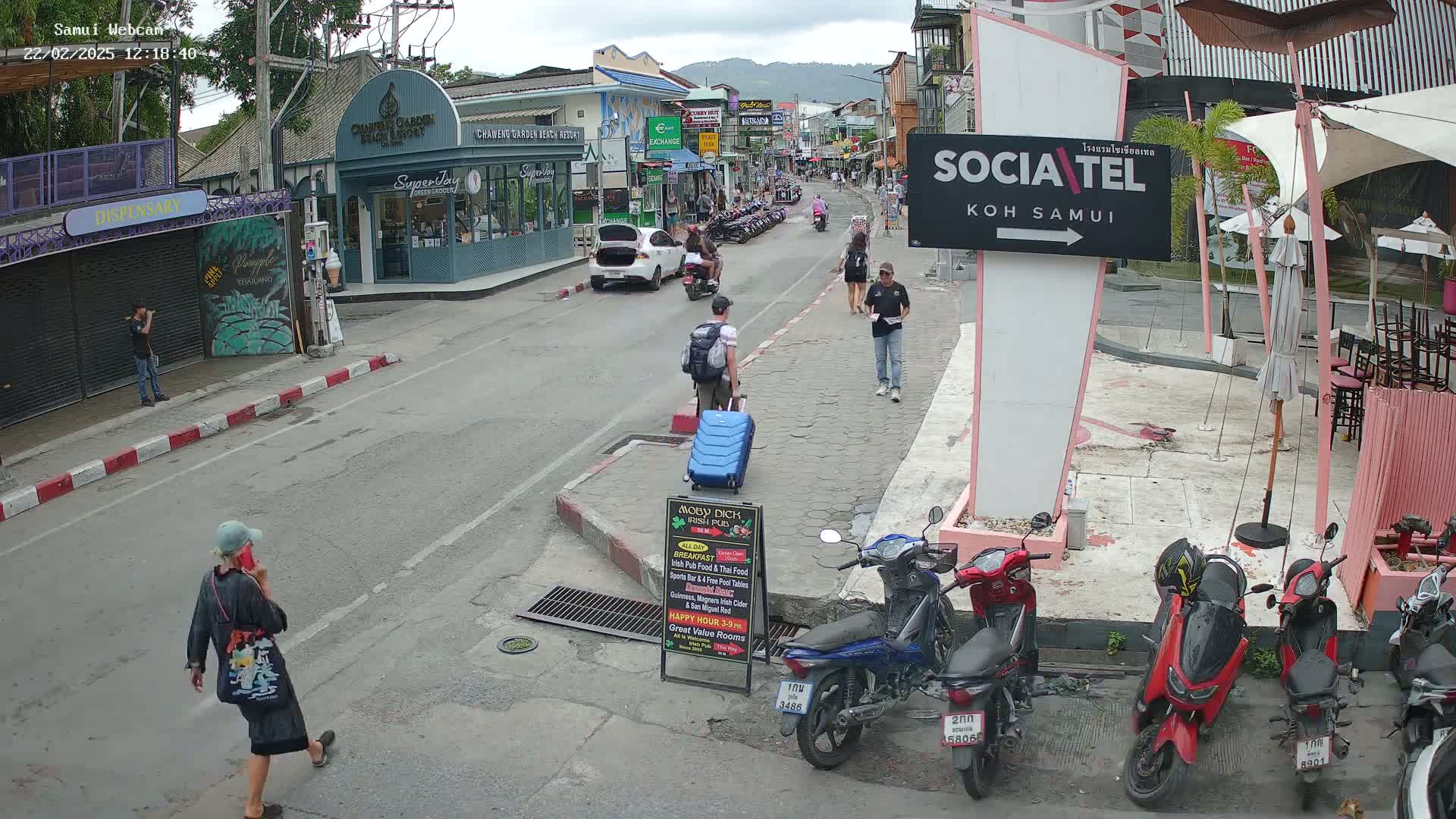 A street scene shows a partly cloudy day with people walking and motorbikes parked along a roadway lined with shops and restaurants.