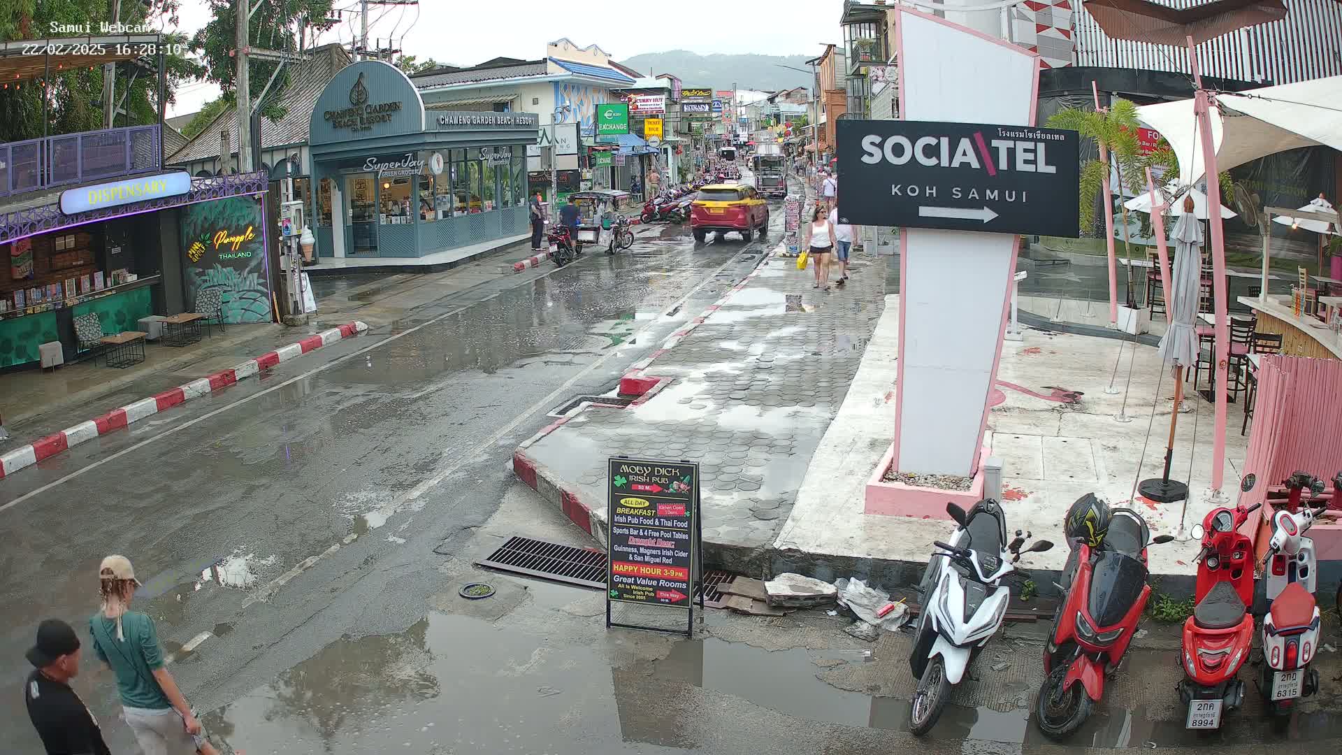 A wet street scene in a town shows several people walking, motorbikes parked, and shops along the sides of the road under an overcast sky.
