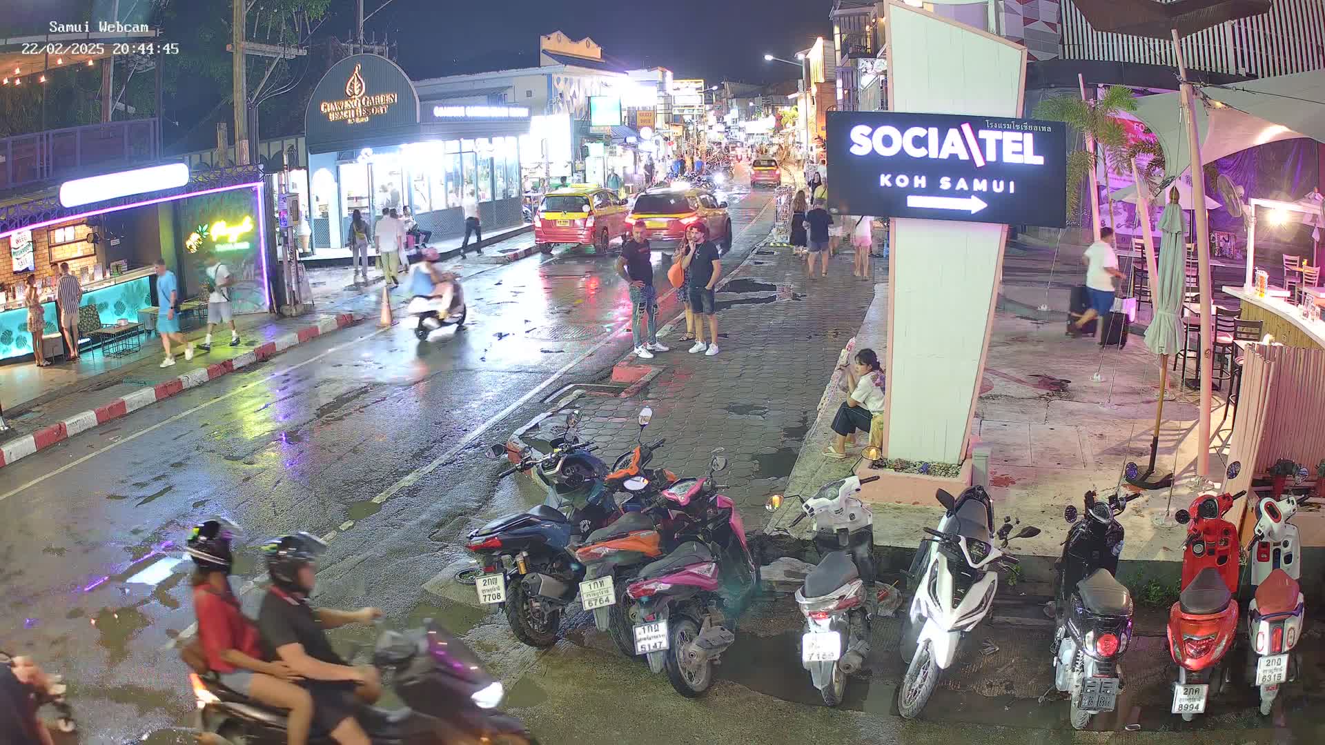 A wet, nighttime street scene shows pedestrians and motorbikes parked along the side of a road lined with shops and businesses, with some people standing in small groups.