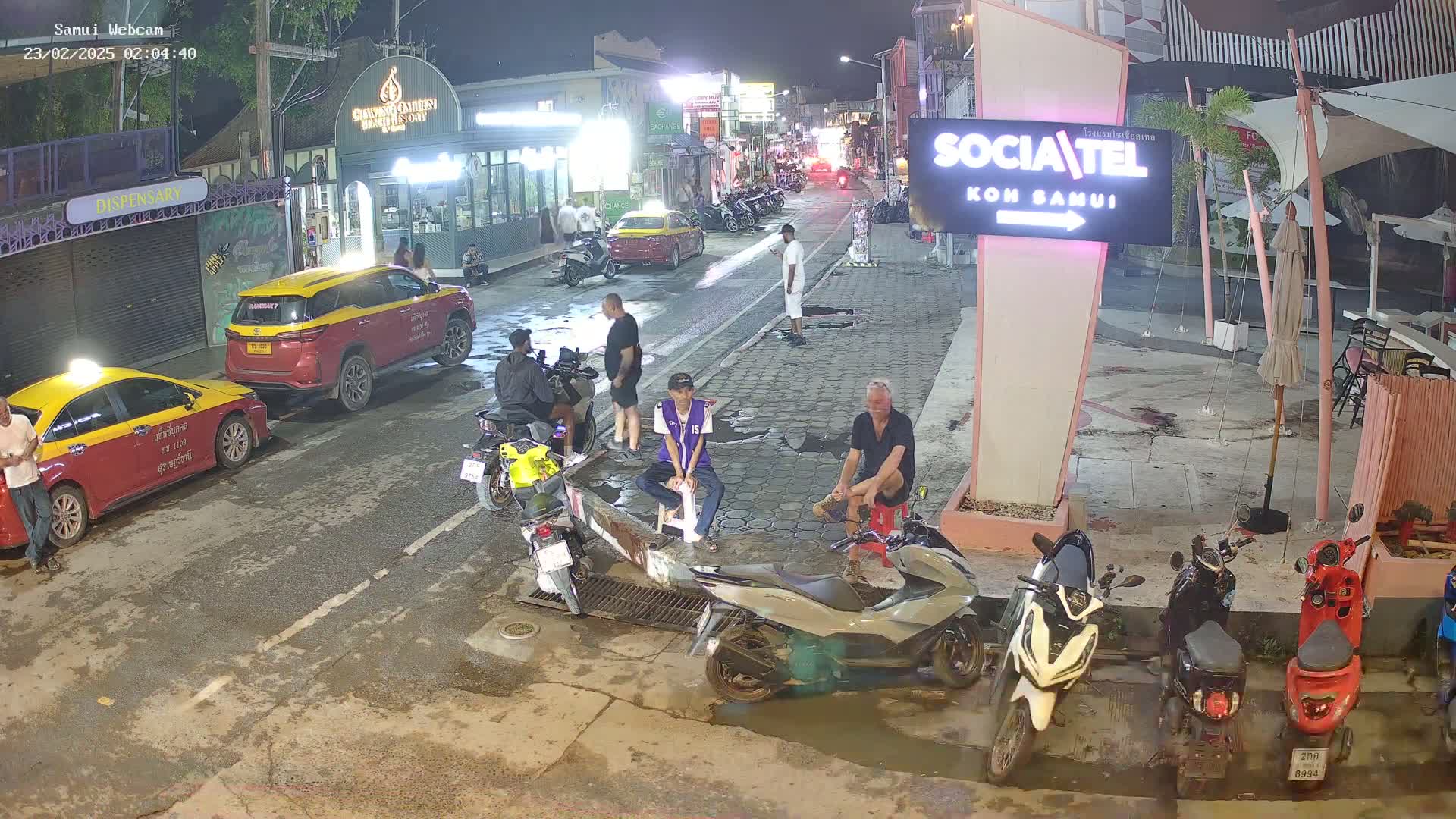 A wet night street scene shows several people, parked taxis, and motorcycles, with some people sitting and others standing around.