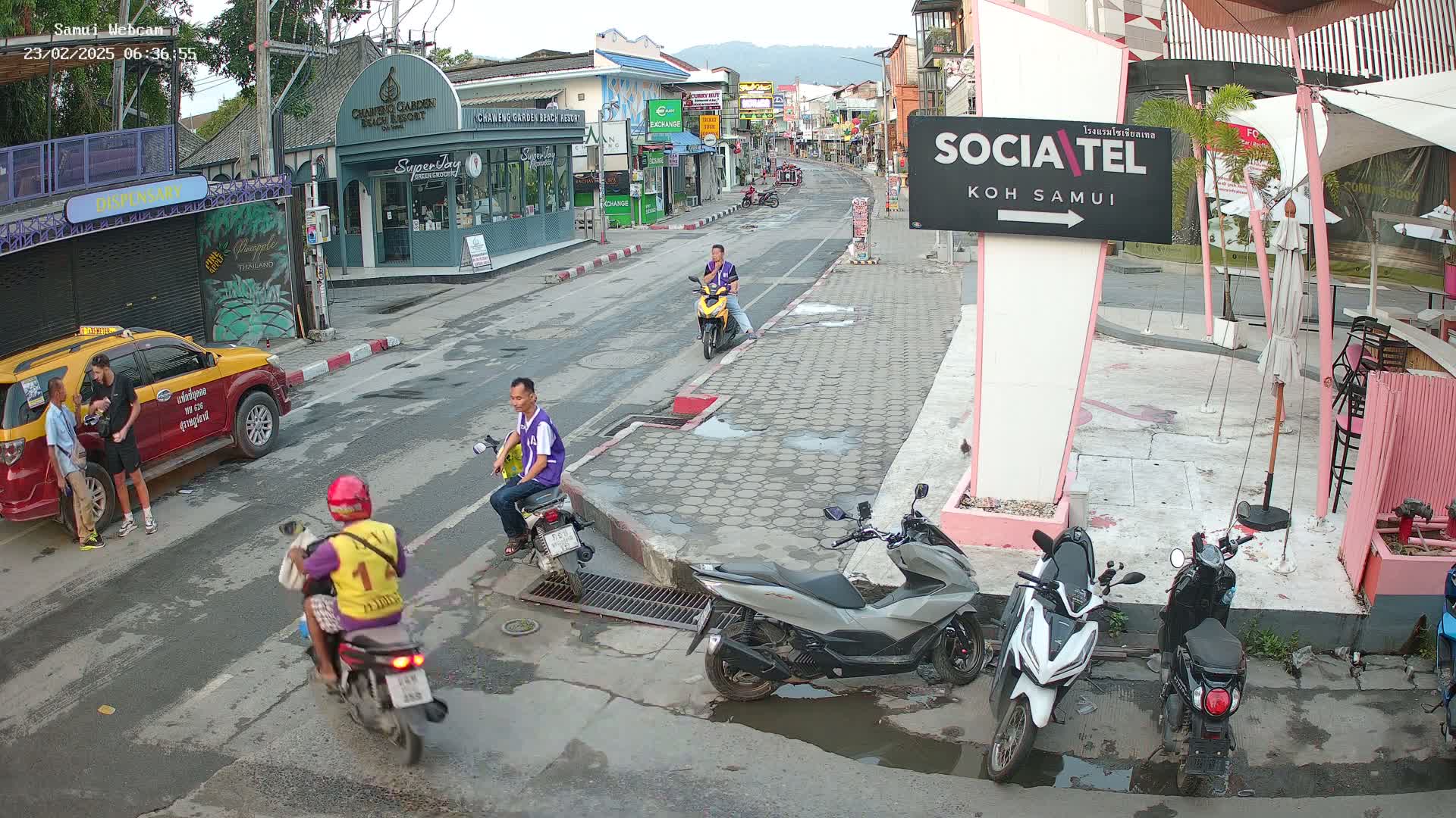 A street scene in Koh Samui, Thailand, shows several people on motor scooters and a taxi, under partly cloudy skies.