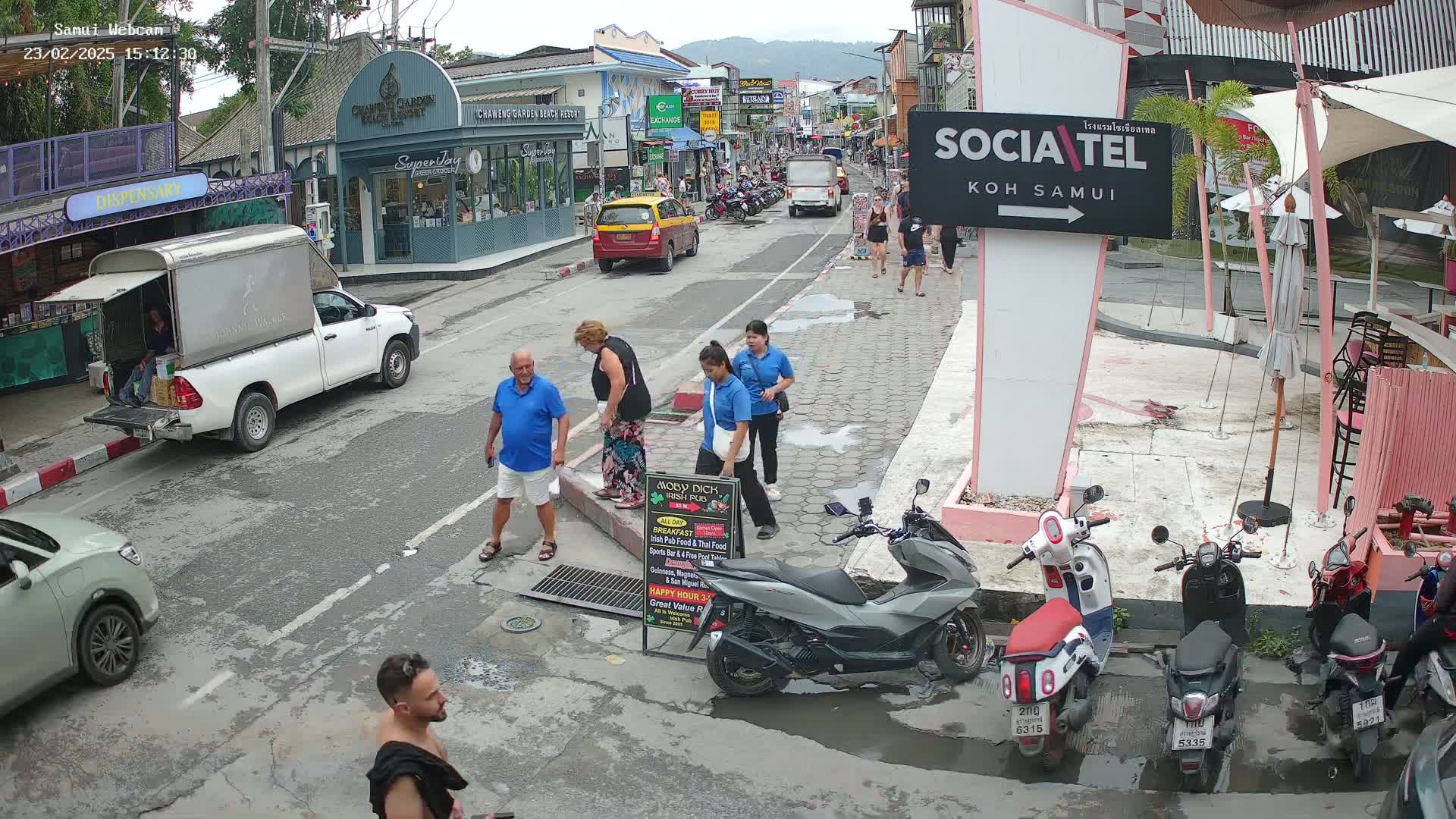 A street scene on a partly cloudy day shows several people walking and standing near parked scooters and cars; a white truck with its tailgate down is parked on the left.