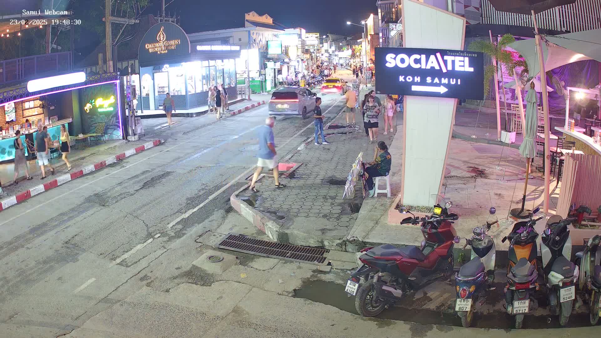 A nighttime street scene shows people walking and standing near shops and restaurants, with several parked motorcycles and cars visible under partly cloudy skies.