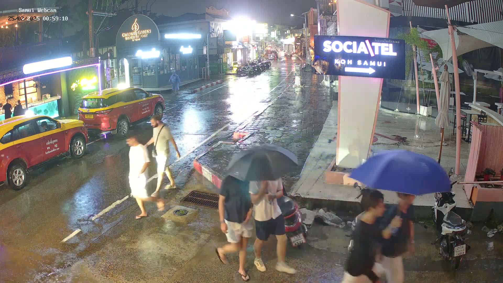 Several people walk along a rain-slicked street at night, some carrying umbrellas, past parked taxis and motorbikes.