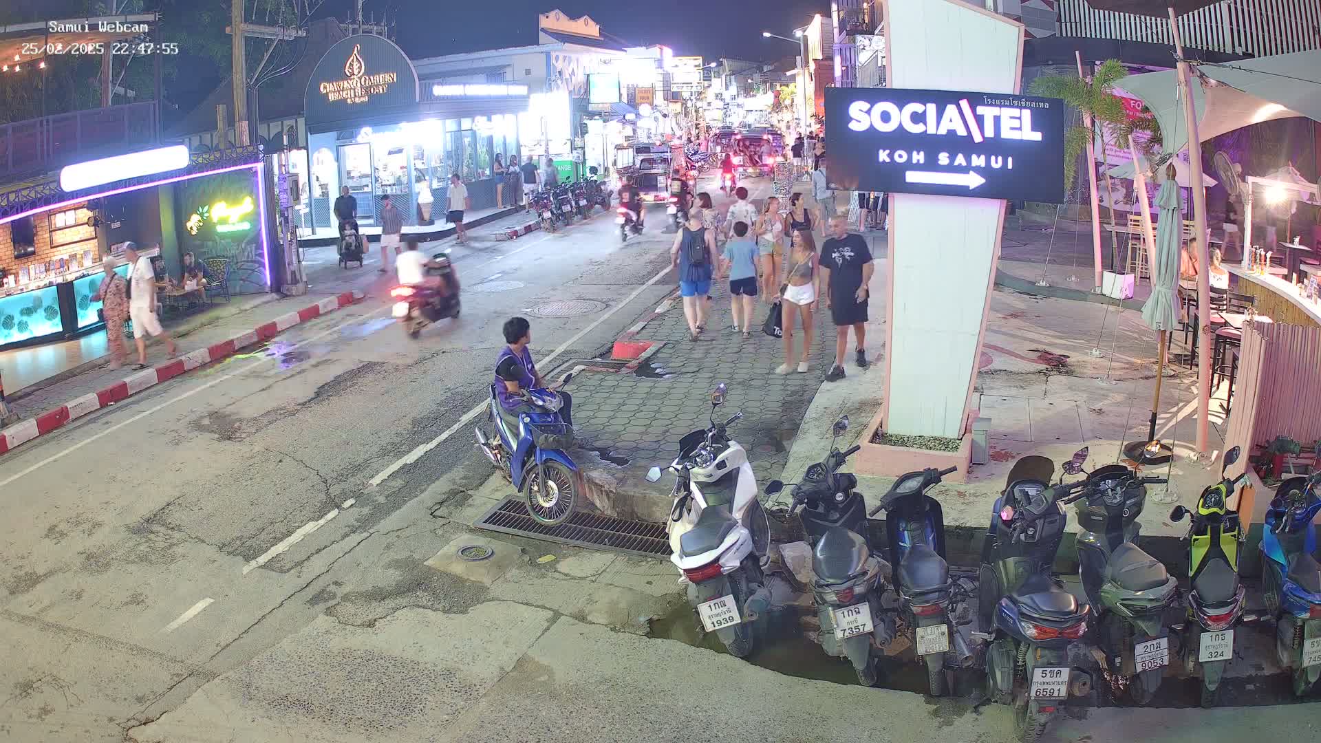At night, a street scene shows people walking and riding motorbikes, with several parked scooters along a curb near shops and restaurants.