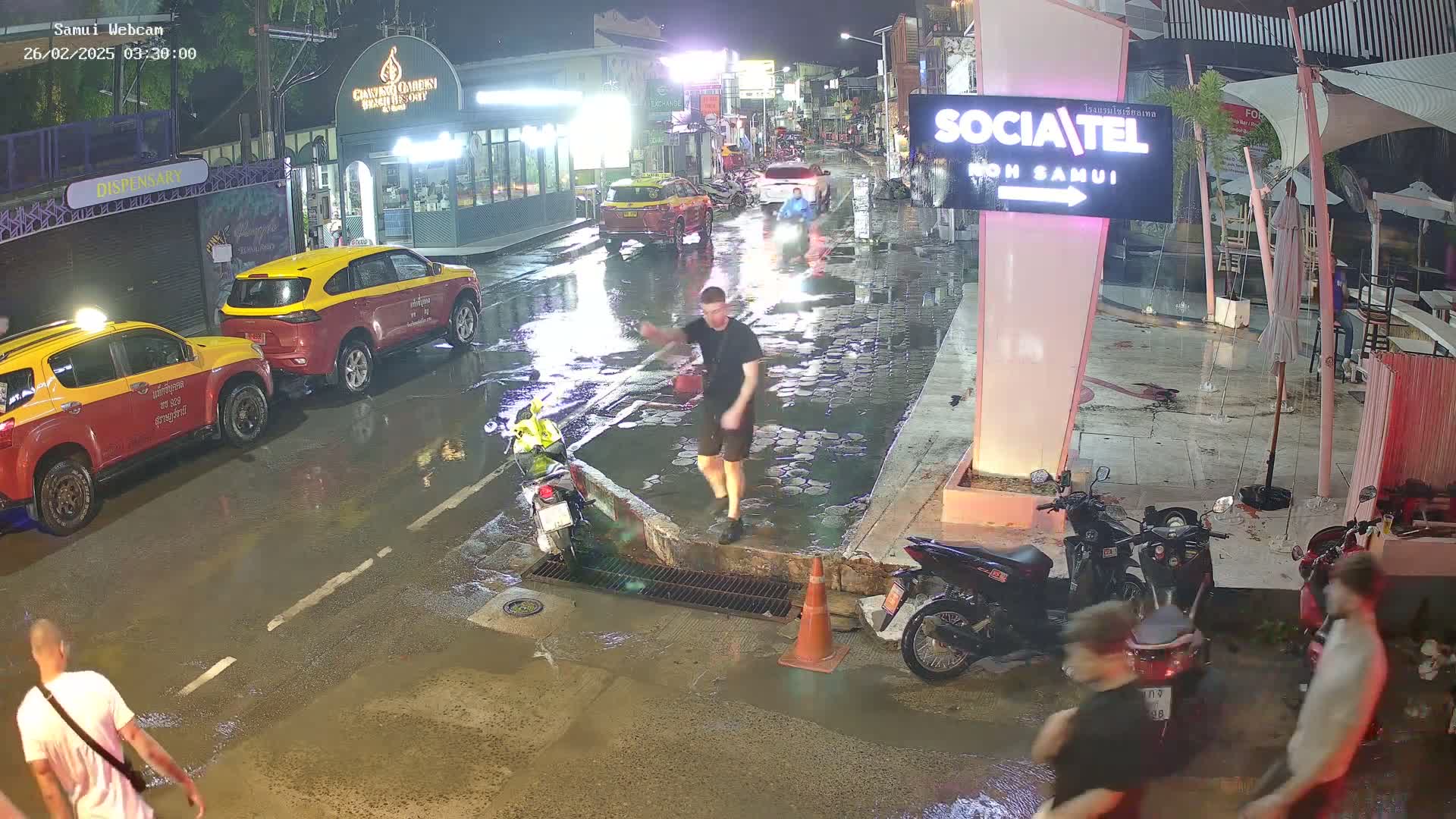 A rain-soaked street at night shows several parked cars and motorbikes, with people walking near a curb and drain.