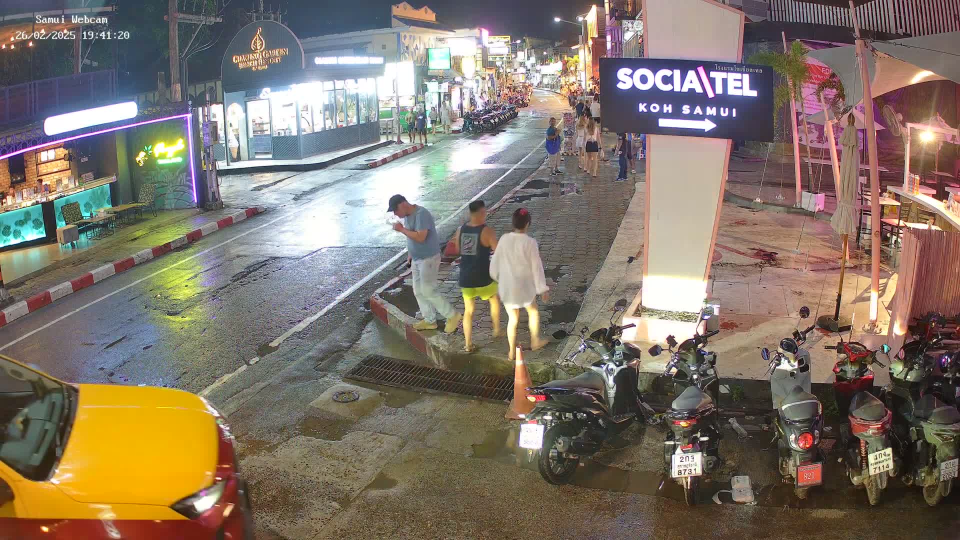 Three people walk on a rain-slicked street at night past parked motorbikes and shops, some with outdoor seating.