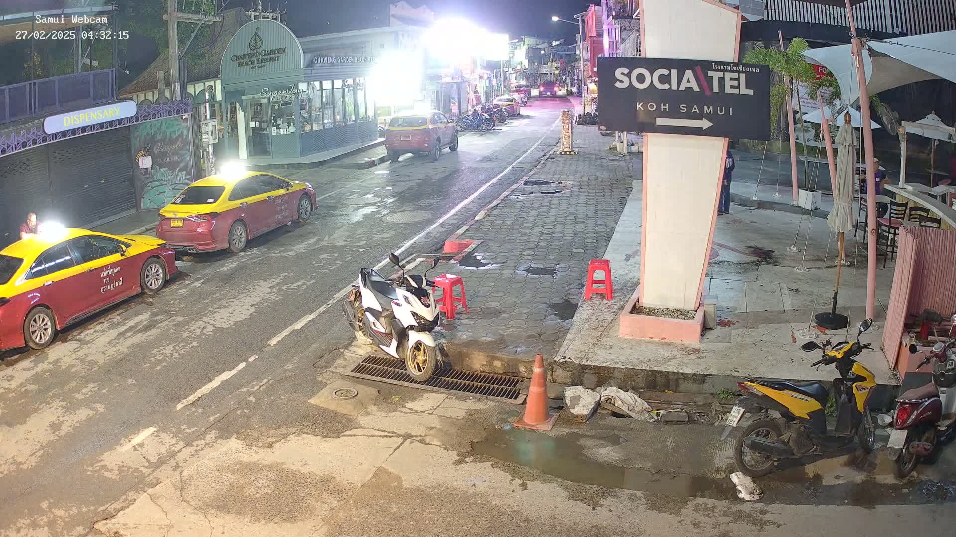At night, a street scene shows two taxis parked near a building, with several motorbikes and scooters parked along the sidewalk, and puddles of water on the street.