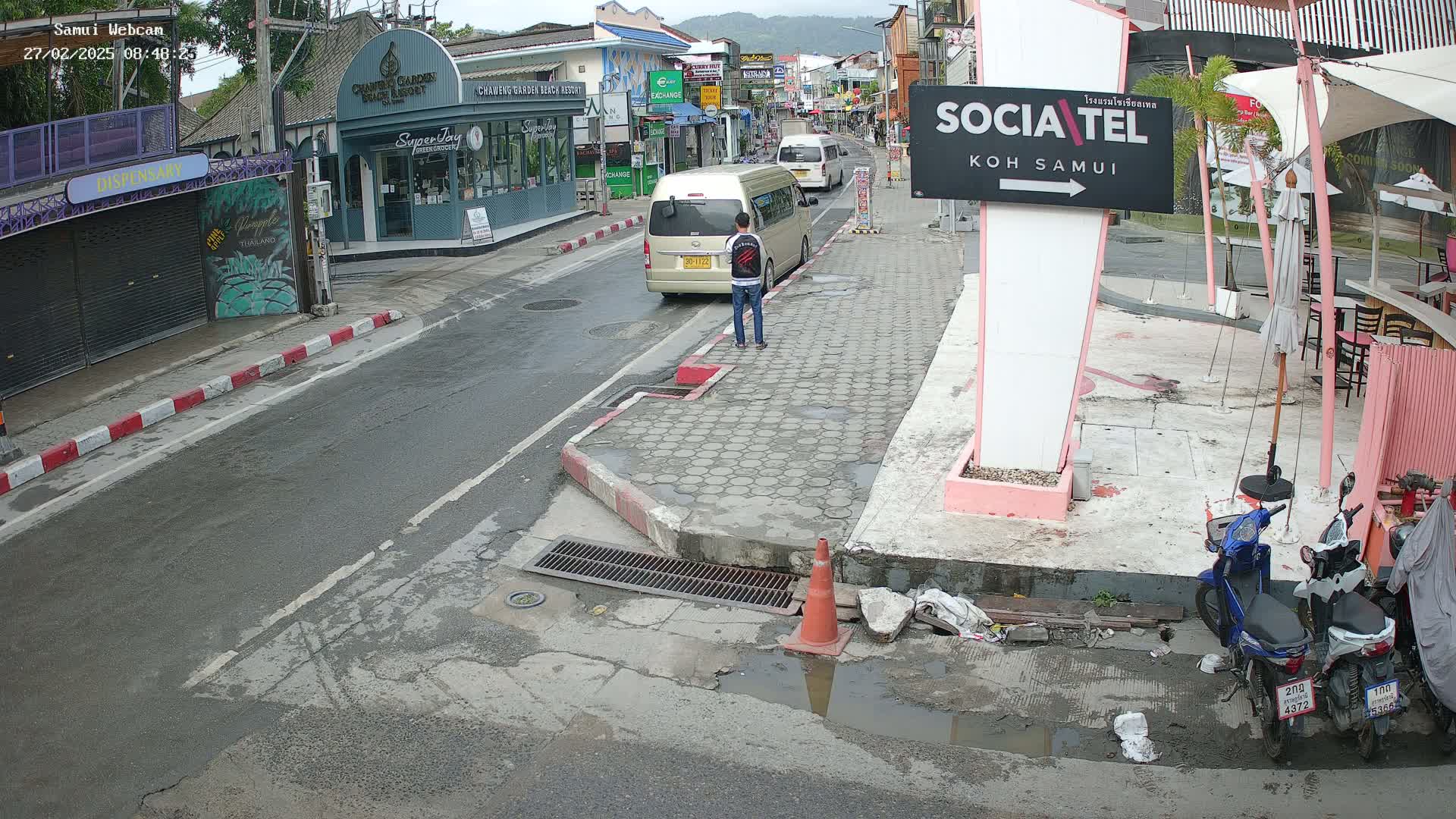 A person stands on a wet, paved street in front of a beige van parked among shops and restaurants, with two motorbikes parked nearby.