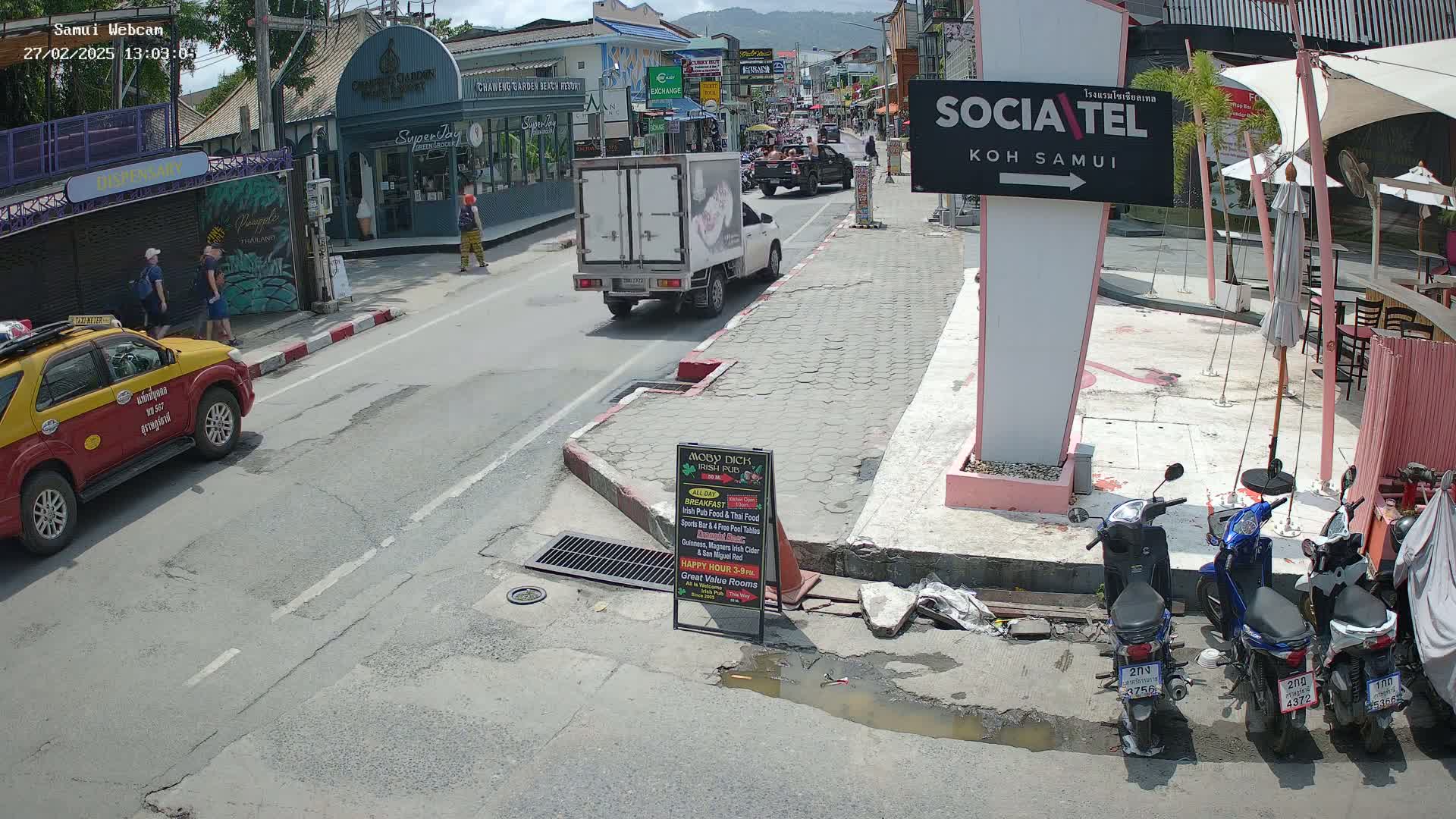 A sunny street scene shows a white delivery truck and a yellow taxi parked near pedestrians and several parked motorbikes, in front of shops and restaurants.