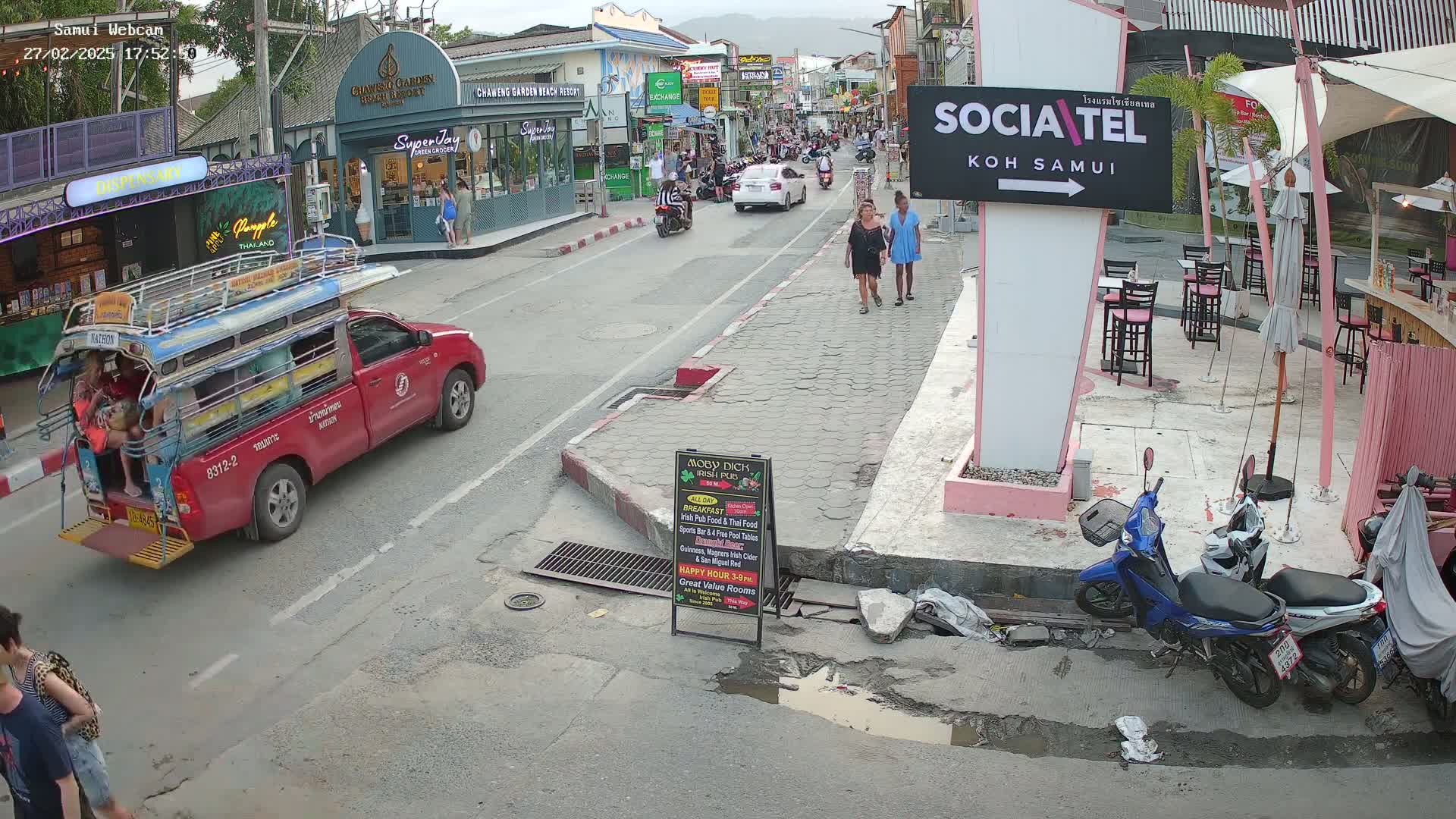 A street scene shows a red songthaew and other vehicles on a road, with pedestrians walking past shops and outdoor seating areas under an overcast sky.