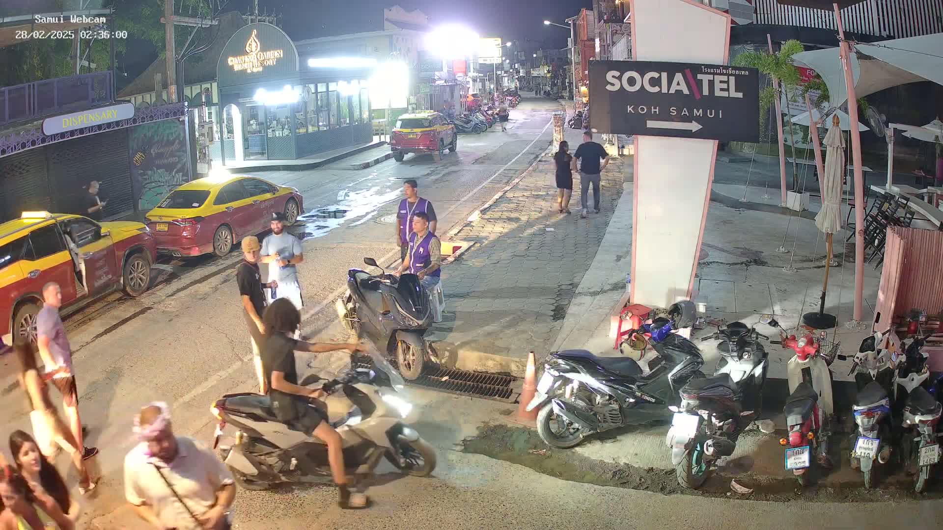 A nighttime street scene shows people standing and sitting around several parked motorcycles and taxis, under clear skies.