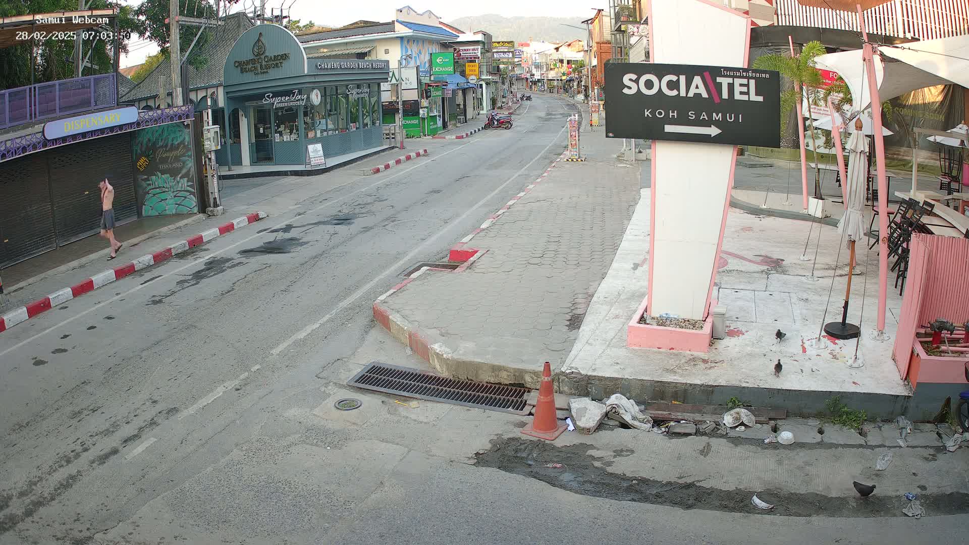 A mostly empty street scene on a sunny day shows a man walking in the distance past shops and restaurants; some litter is visible on the ground near a pink sign.