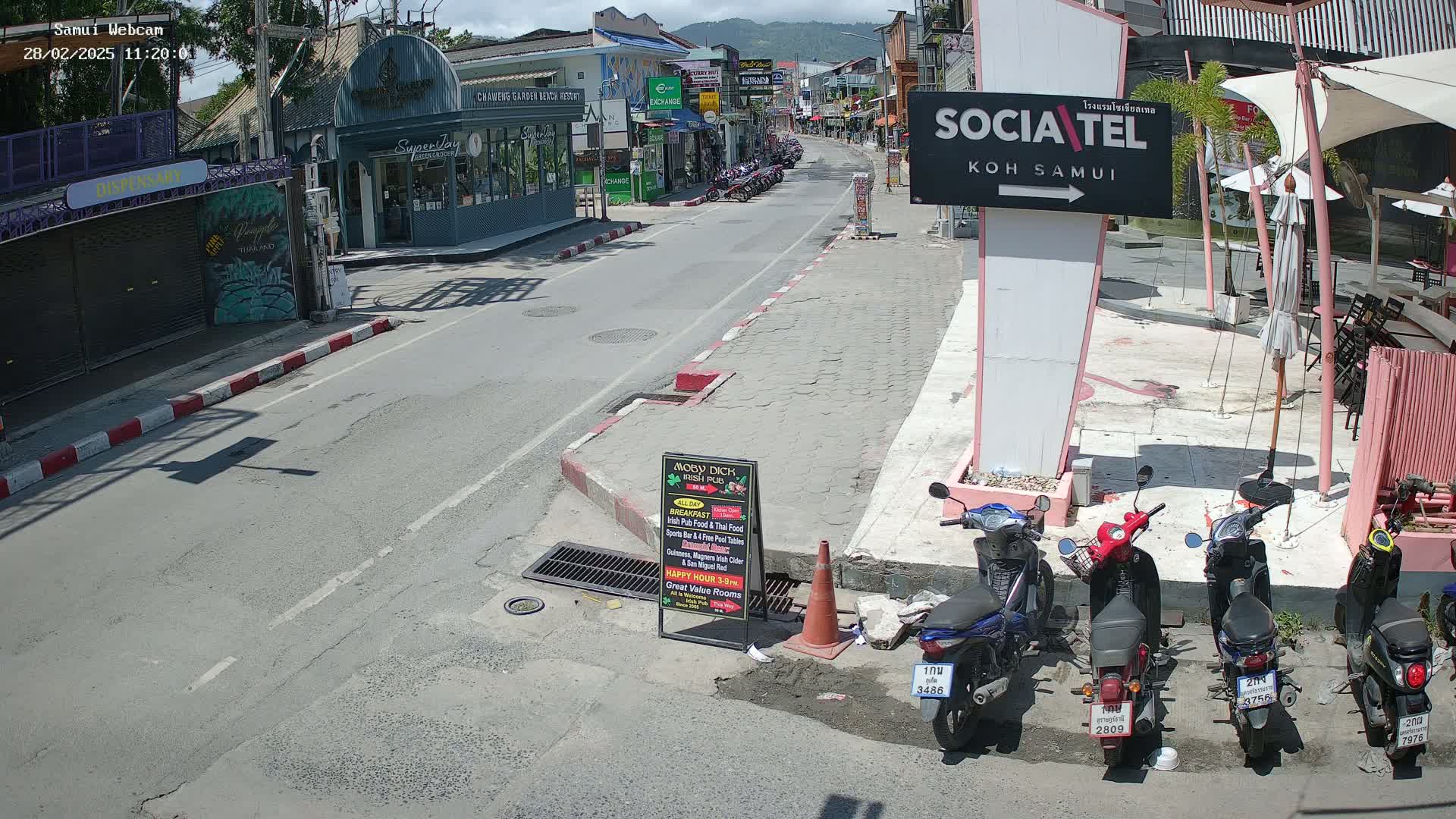 A sunny street scene shows several parked motorbikes alongside a roadway lined with shops and restaurants, under a mostly clear sky.