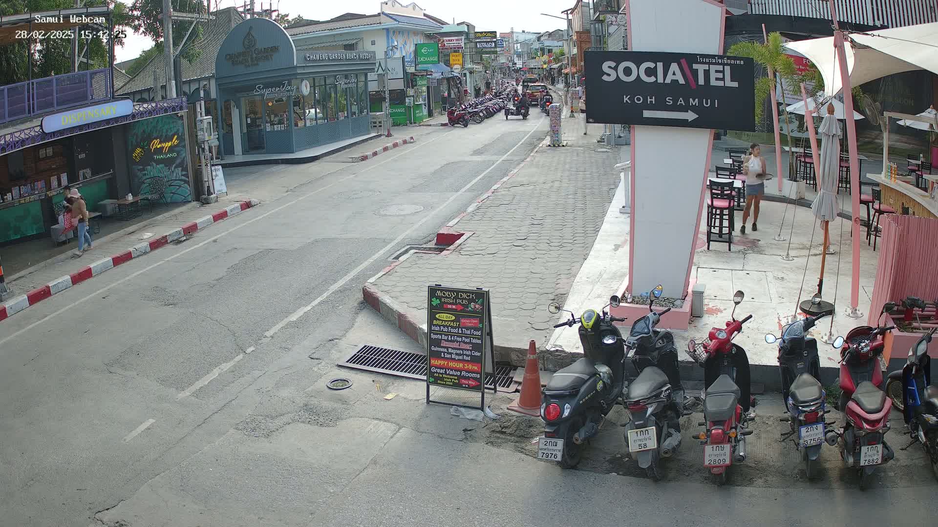 A street scene shows parked motorbikes lining the sides of a road, with pedestrians and shops along the sides; the weather is sunny.