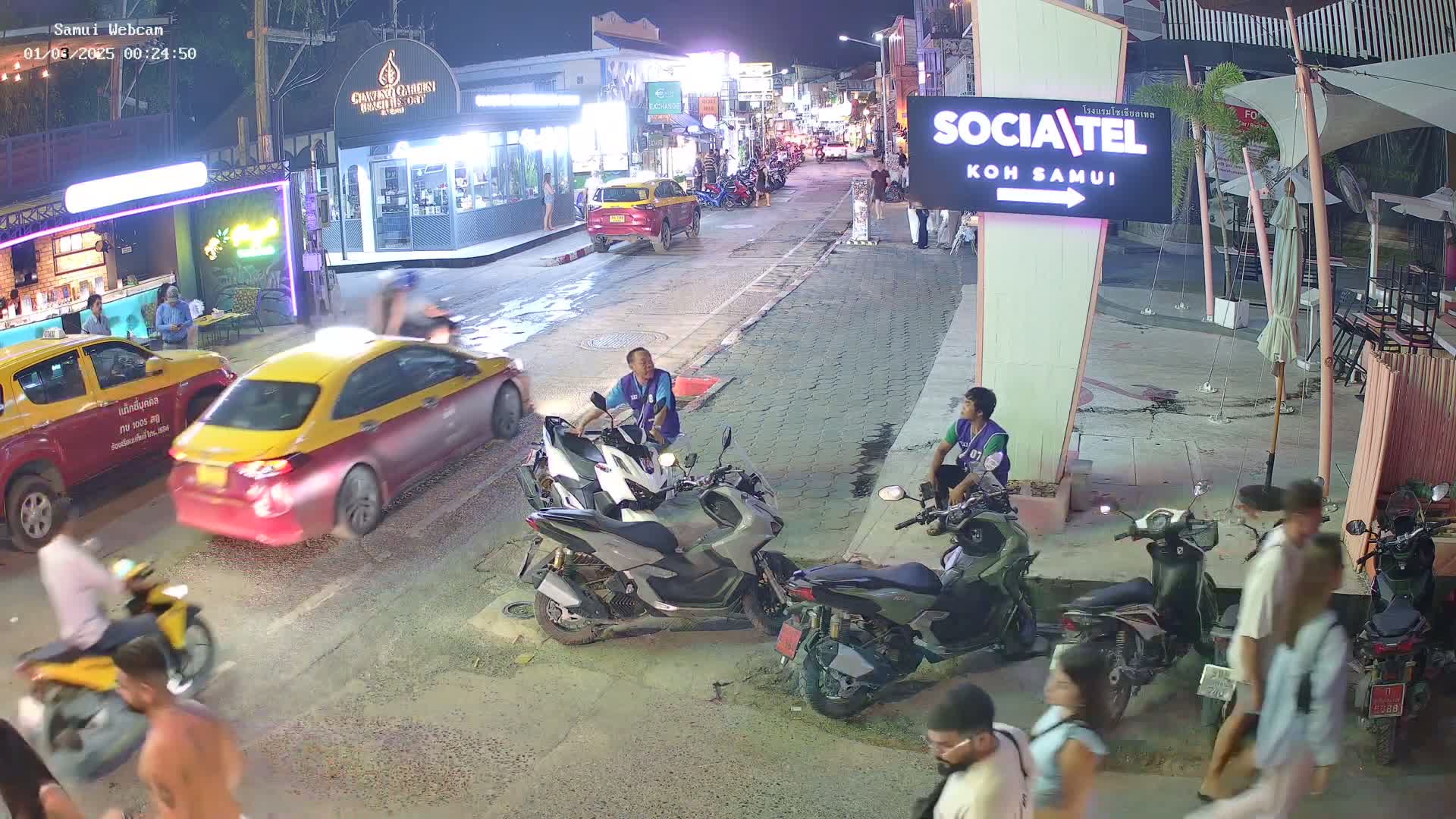 At night, a street scene shows several people walking and standing near parked motorcycles and taxis, under a clear sky.