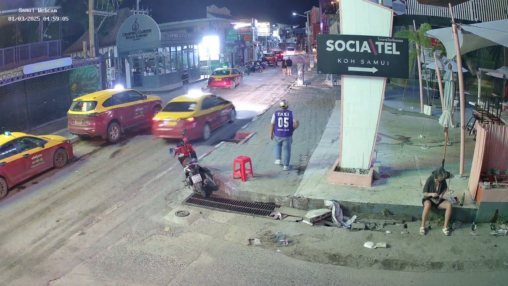 At night, several taxis are parked or driving on a street near a building, with a person sitting on the ground nearby.