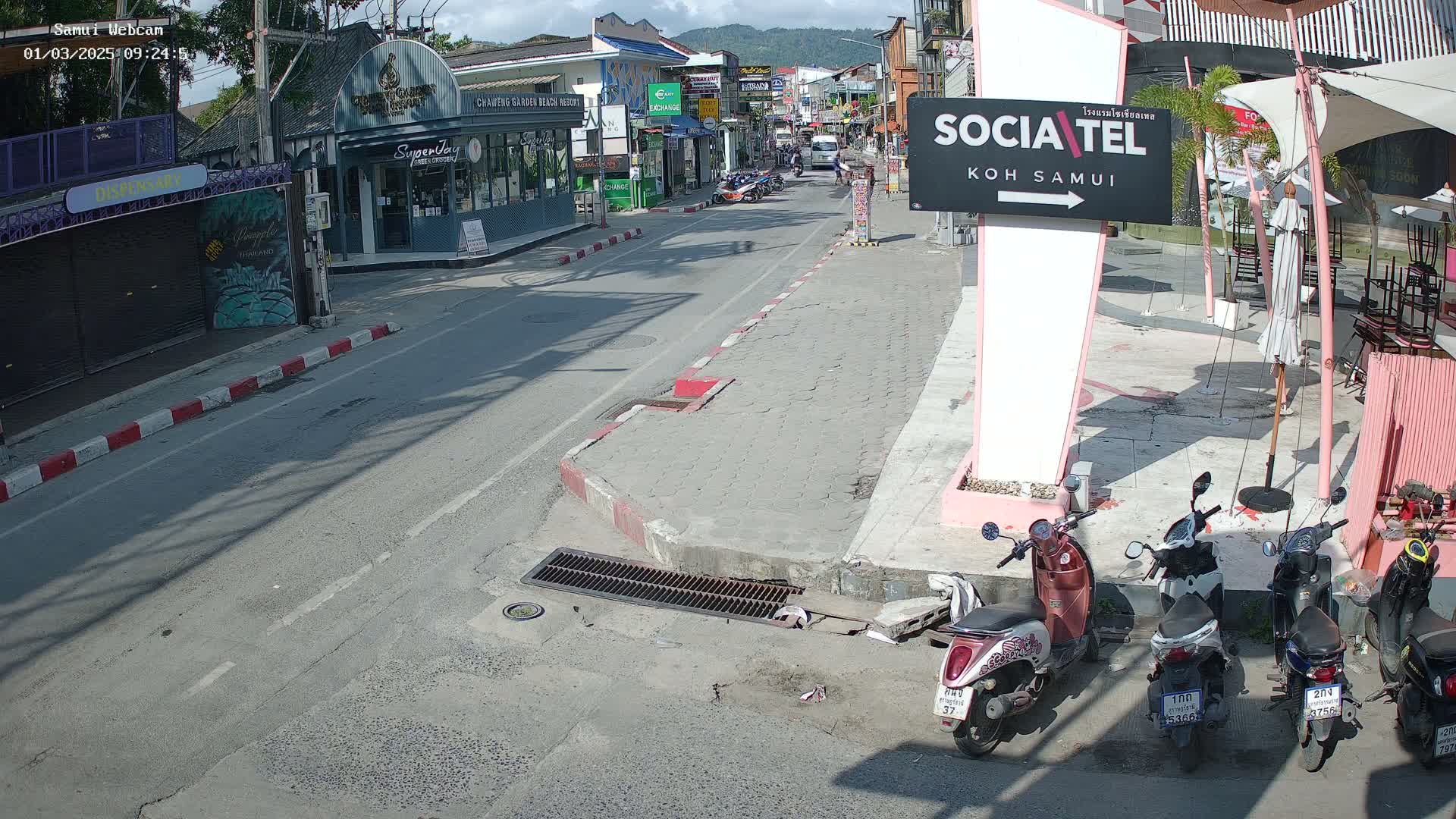 A sunny street scene shows a road with parked motorbikes near shops and restaurants.