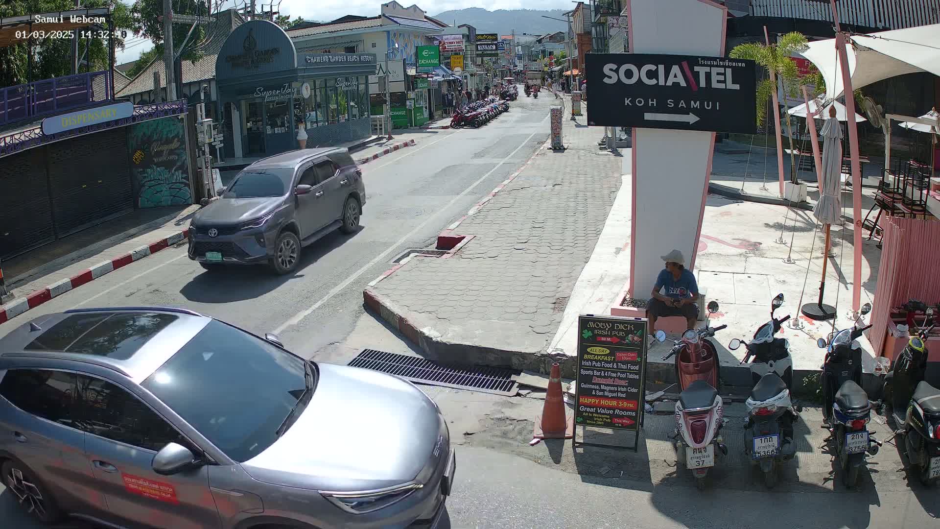 A sunny street scene shows parked cars and motorbikes, along with a few pedestrians near shops and restaurants.