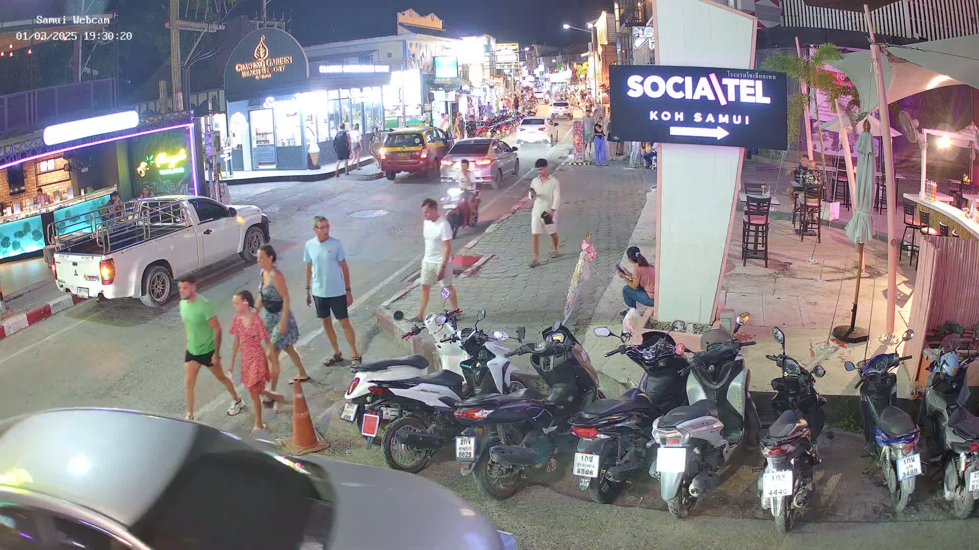 At night, several people walk along a street lined with shops and restaurants, while numerous motorbikes are parked nearby.