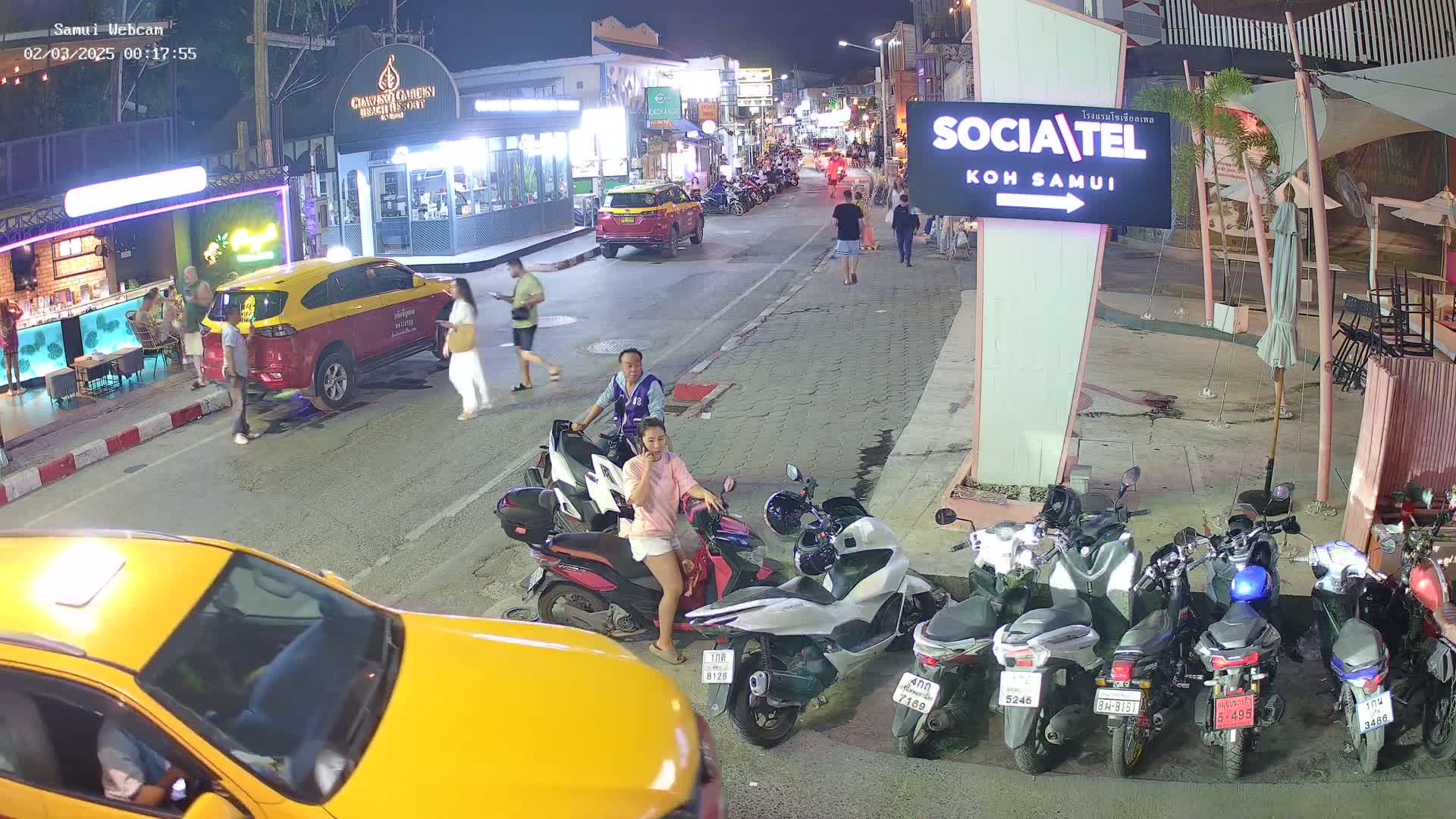 At night, a street scene shows people walking, taxis and motorbikes parked, and several businesses along the side of the road.